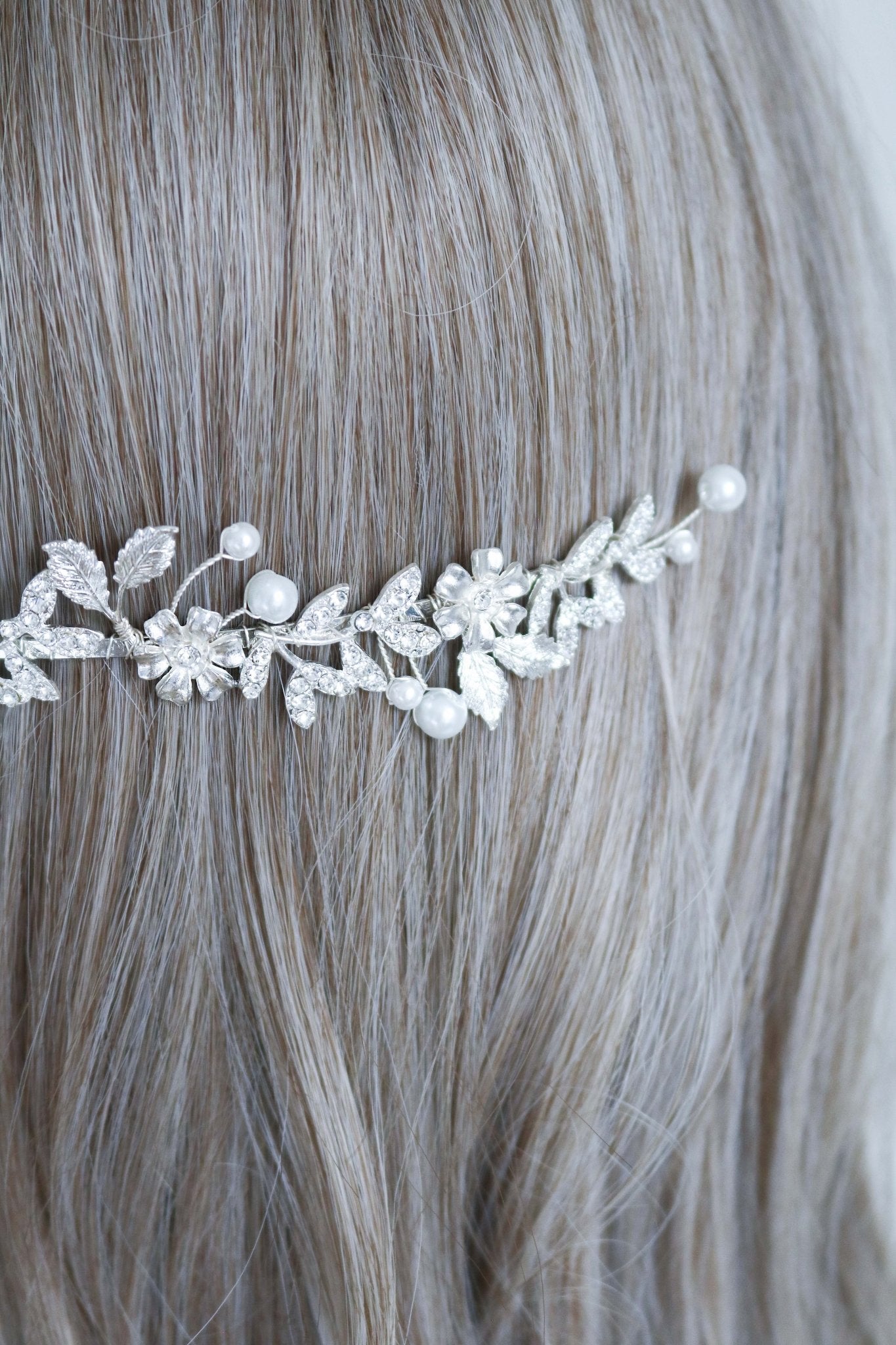 A close up of a silver wedding headband with crystal leaves, flowers, and freshwater pearls sits behind a head of hair. 