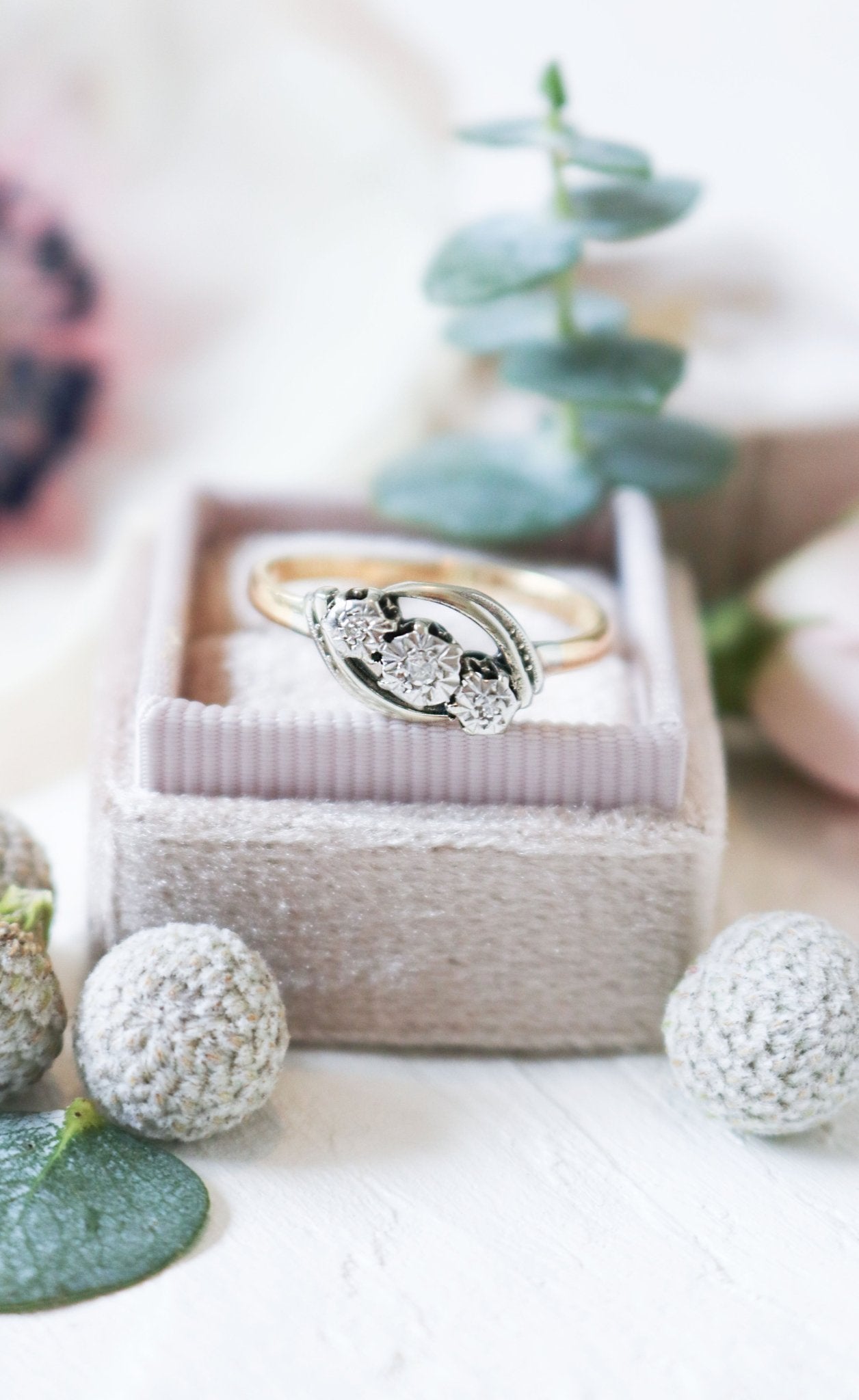 A close up of a vintage engagement ring in gold and platinum with diamonds sits in a ring box surrounded by flora. 