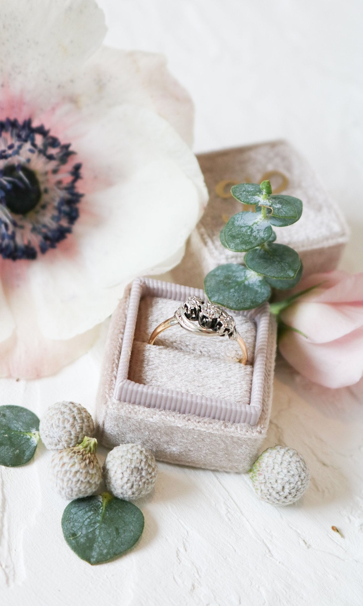 A vintage engagement ring in gold and platinum with diamonds sits in a ring box surrounded by flora. 