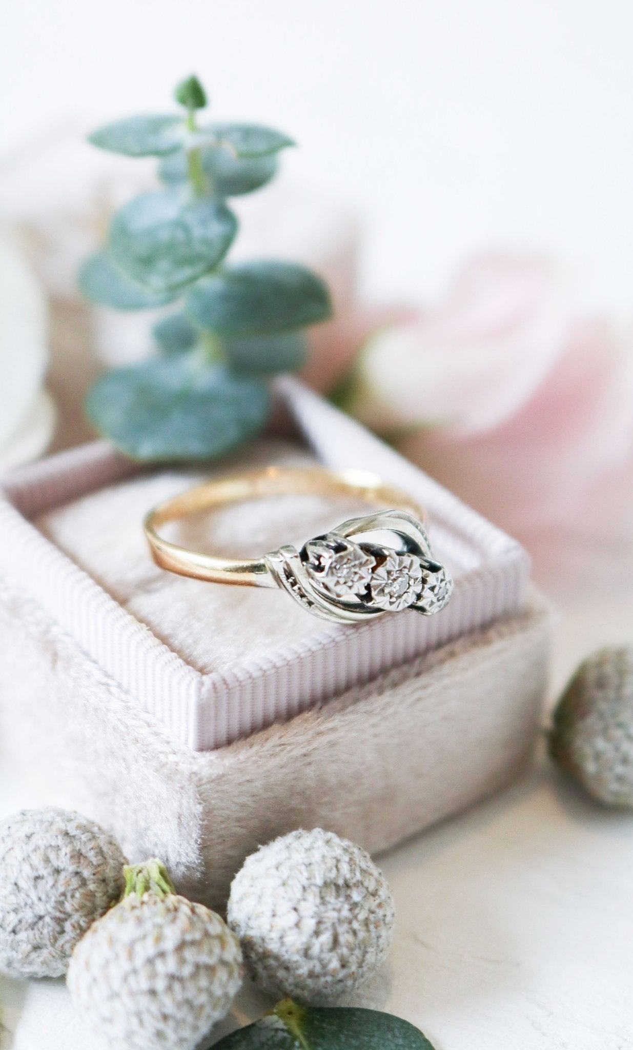 A vintage engagement ring in gold and platinum with diamonds sits in a ring box surrounded by flora. 