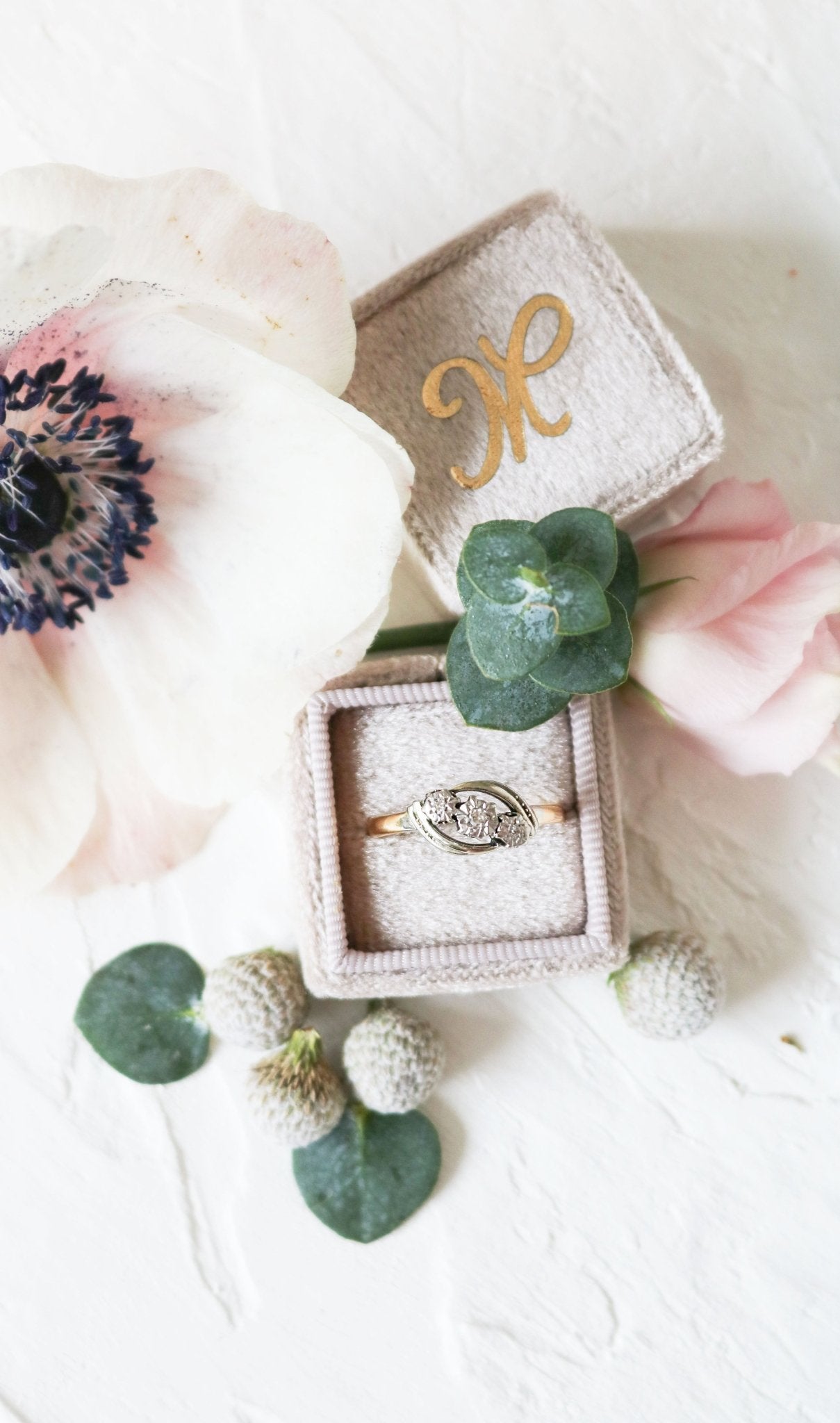 A vintage engagement ring in gold and platinum with diamonds sits in a ring box surrounded by flora. 