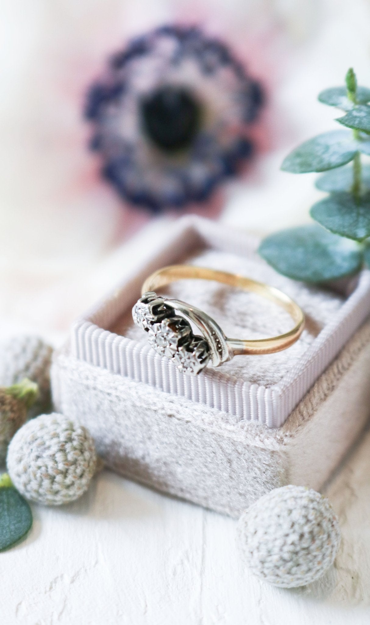 A close up of a vintage engagement ring in gold and platinum with diamonds sits in a ring box surrounded by flora.