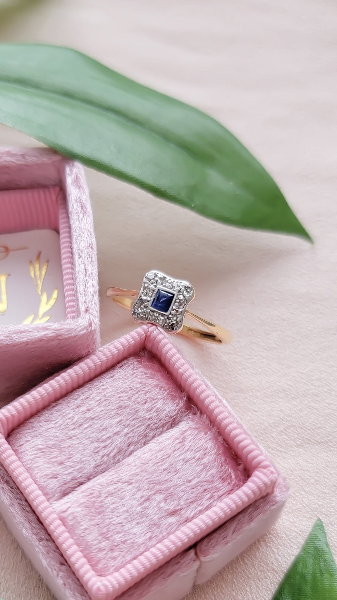 A vintage engagement ring in yellow gold with platinum, diamonds, and blue sapphire leaning against a pink ring box. 