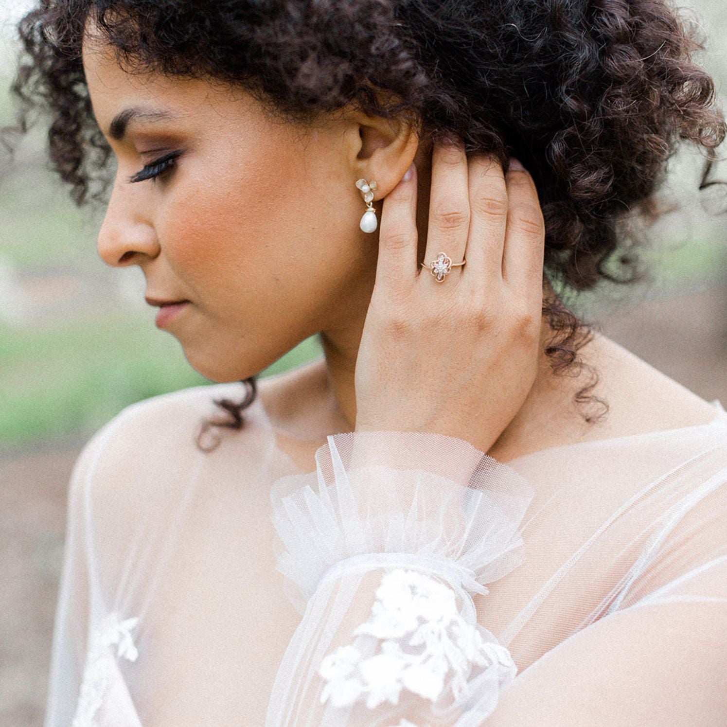A BIPOC bride wearing floral freshwater pearl bridal earrings and a vintage engagement ring, with a blurred background