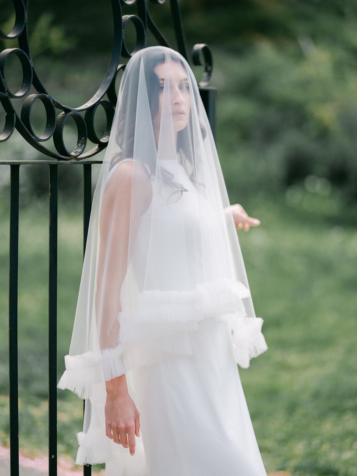 A long, ruffled wedding veil covers a new bride as she stands in front of a cast iron gate. 