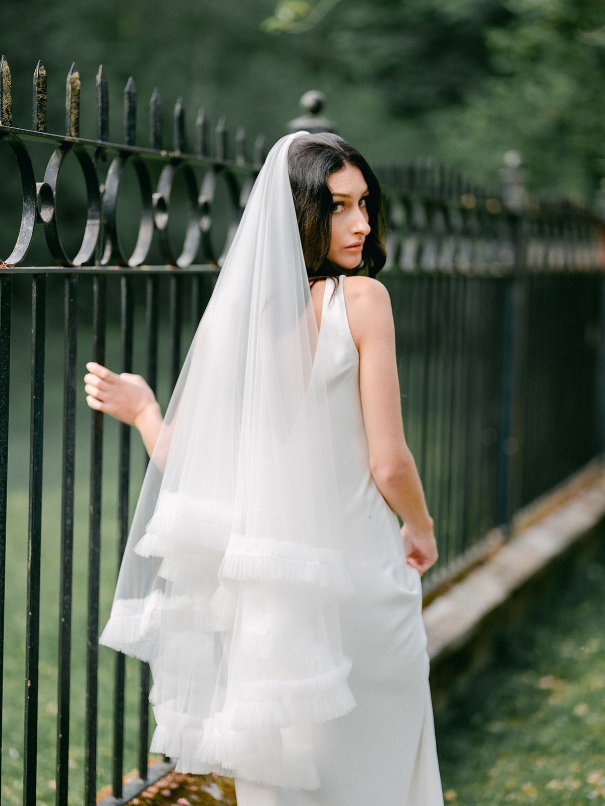 A ruffled wedding veil trails behind a bride looking back and holding on to a cast iron fence. 