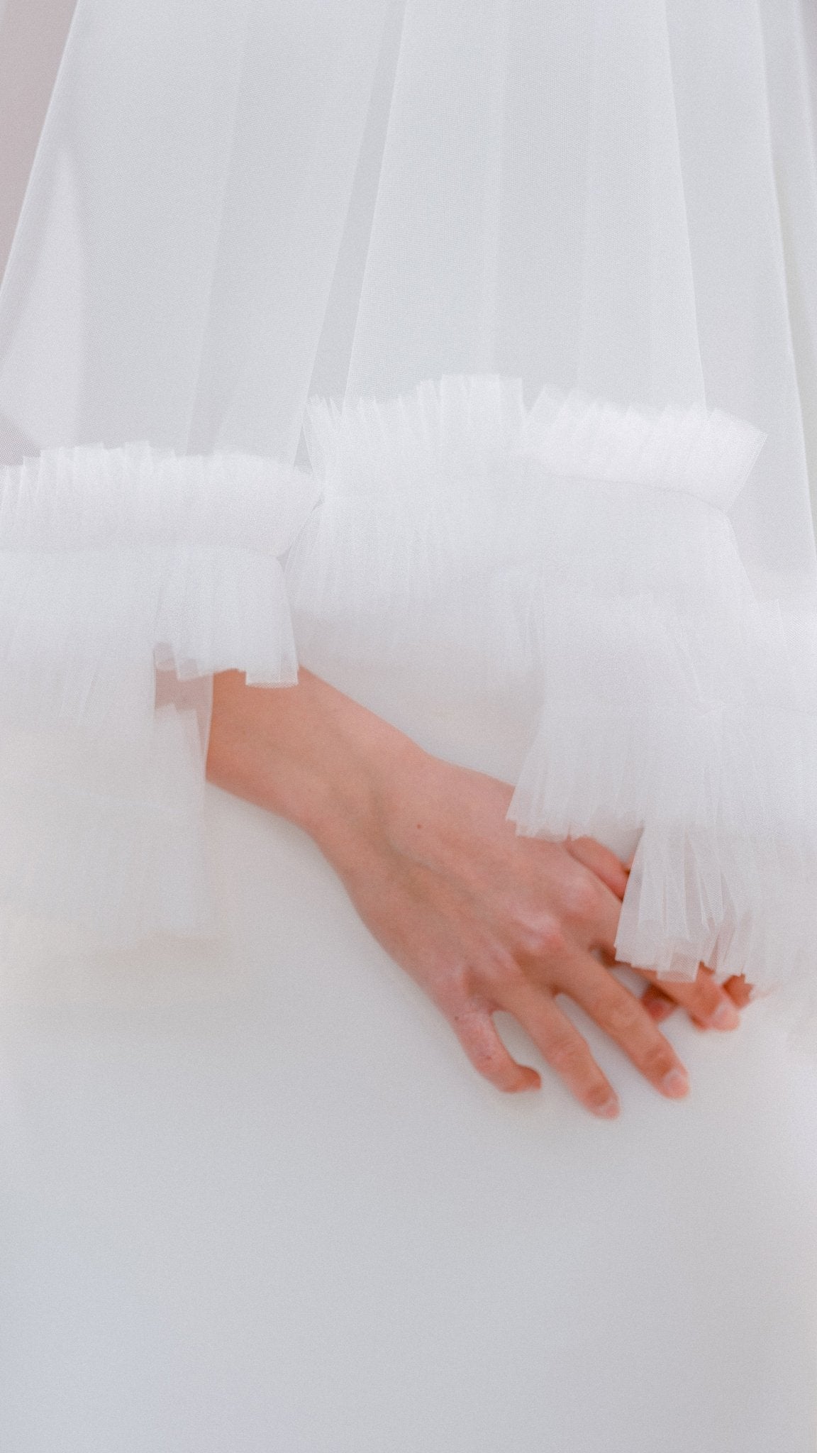 A close up of hands crossed on top of a bridal gown partly covered by a ruffled wedding veil. 