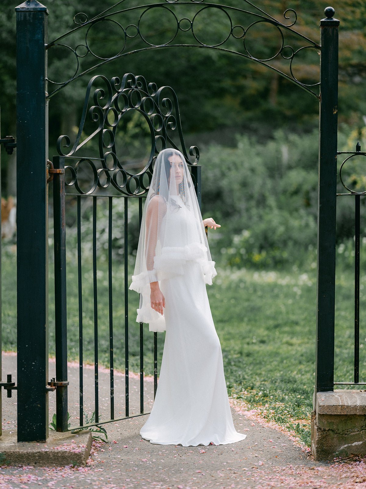 A long, ruffled wedding veil covers a new bride as she stands in front of an open cast iron gate. 