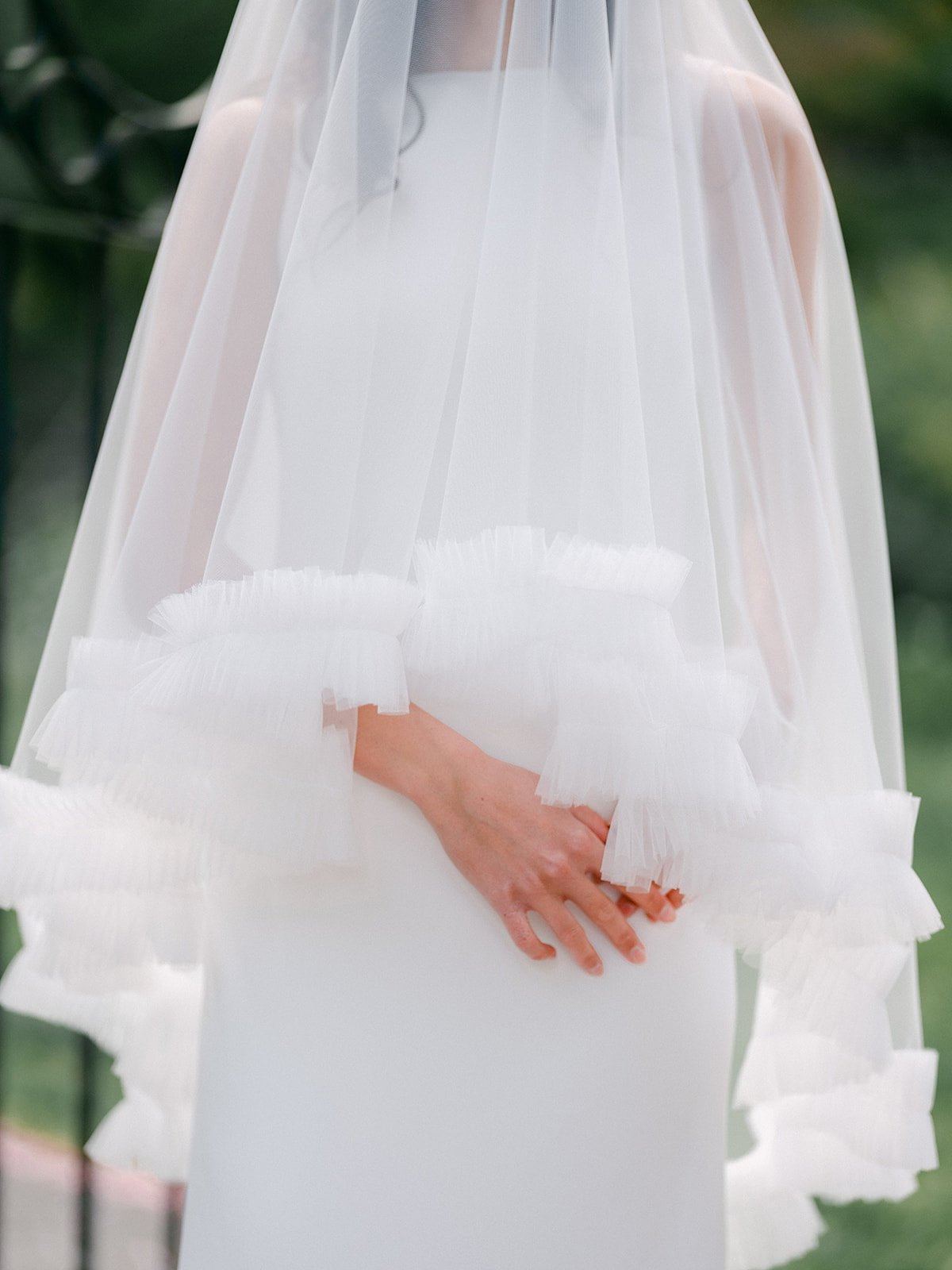 Hands crossed on top of a bridal gown partly covered by a ruffled wedding veil. 