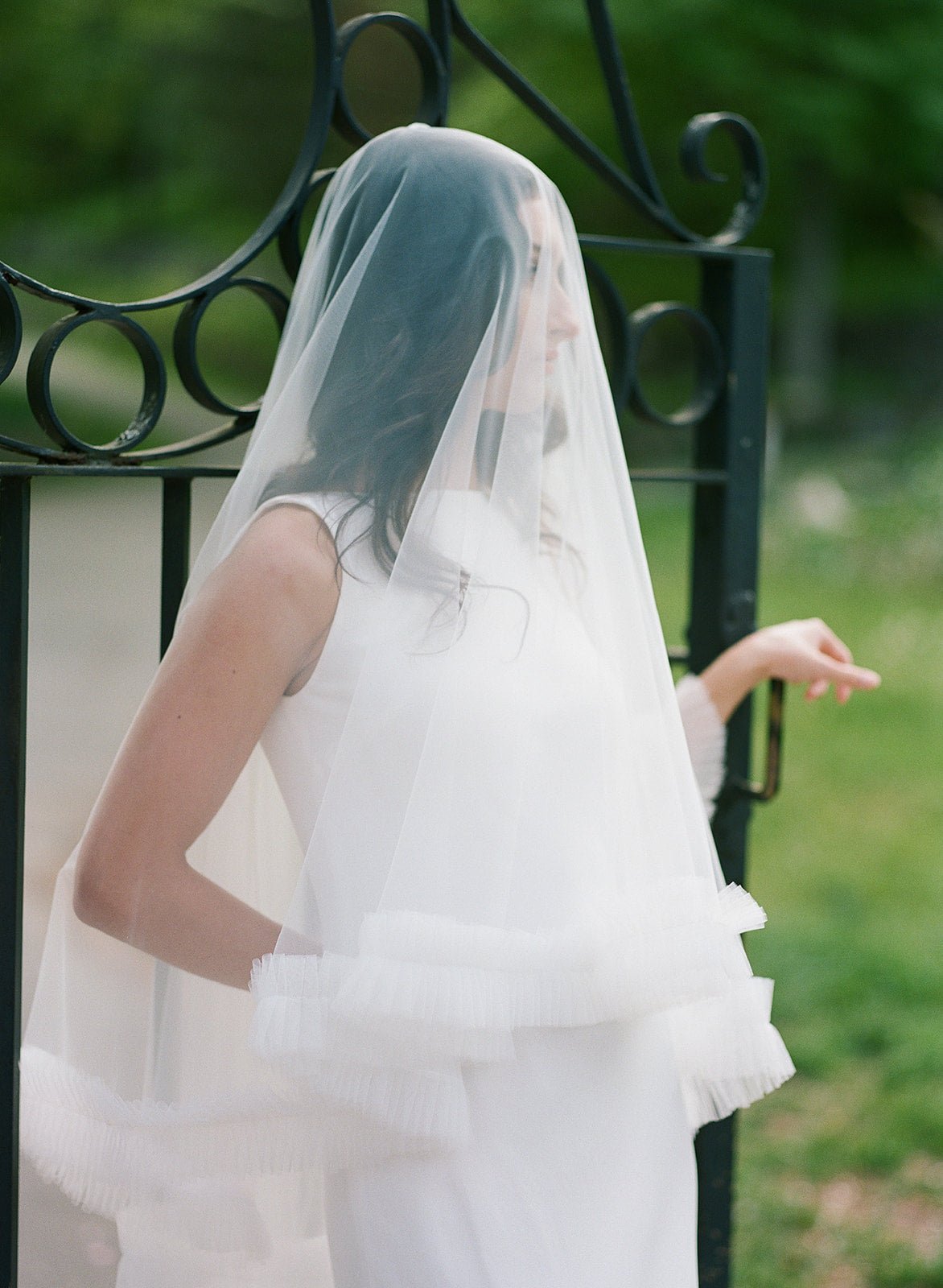 A long, ruffled wedding veil covers a new bride as she looks off to the side and stands in front of an open cast iron gate. 