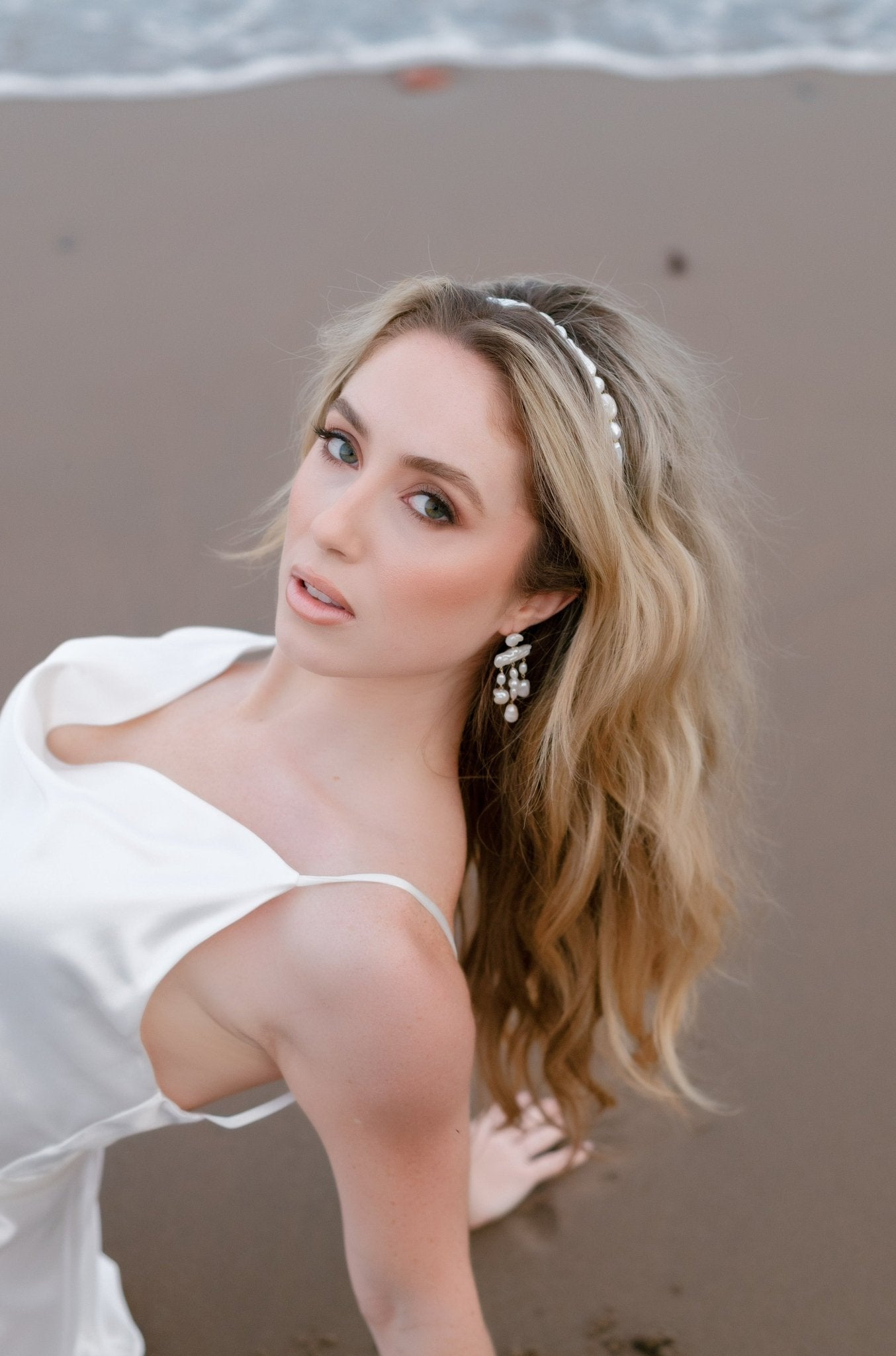 A beautiful bride leans back on a beach in a bridal gown wearing a pearl headband and dangle freshwater pearl earrings.