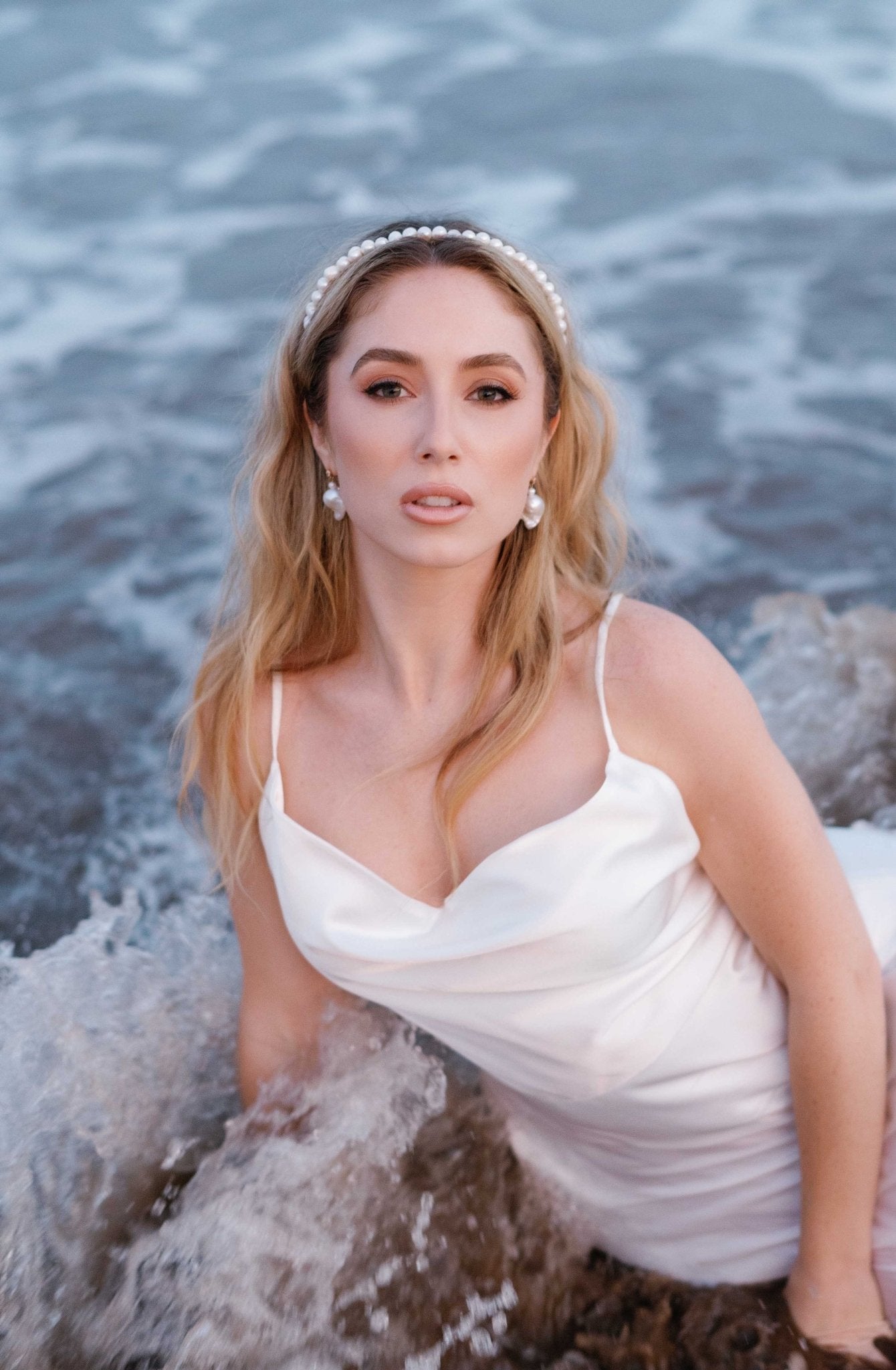 A bride lays among the waves adorned with Baroque freshwater pearl earrings, a bridal pearl headband, and a wedding dress.