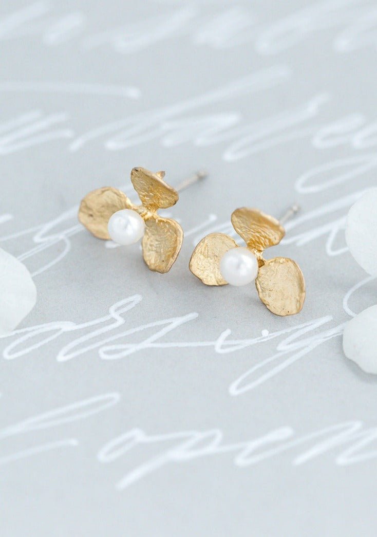 A pair of gold-filled flower stud earrings with organic freshwater pearls and sterling silver posts on a grey surface with writing. Minimalist bridal jewelry & everyday jewelry handmade by Canadian jewelry designer