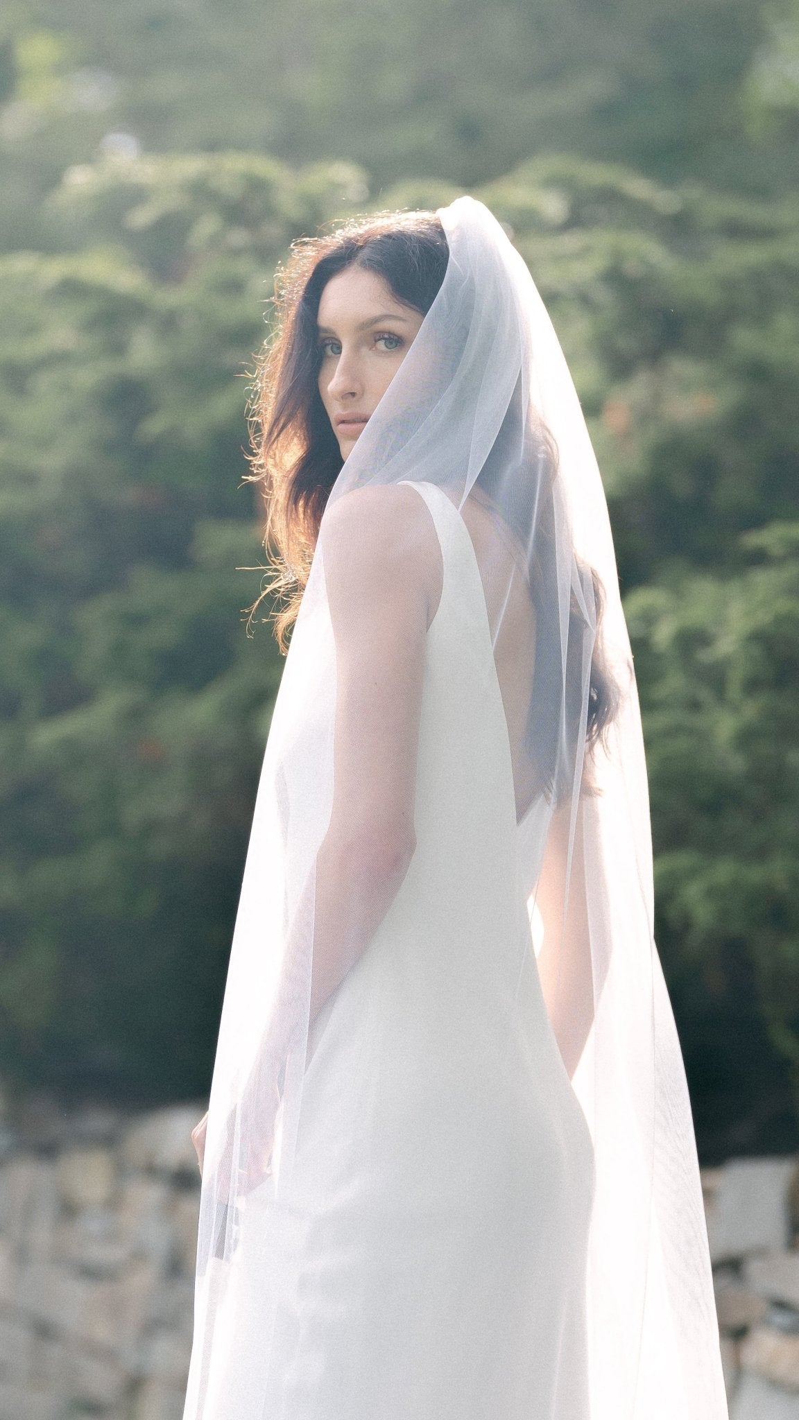 Beloved Traditional Tulle Wedding Veil draped over a bride as she looks over her shoulder with nature background. Wedding veils & bridal hair accessories for Canadian weddings, handmade by affordable Canadian jewelry brand