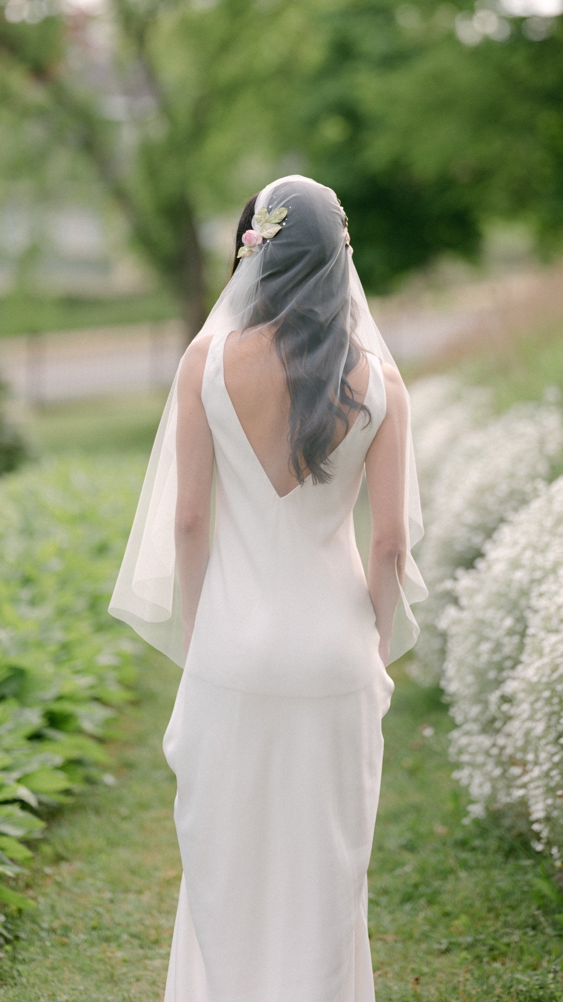 A bride with wedding veil from the back surrounded by greenery. 