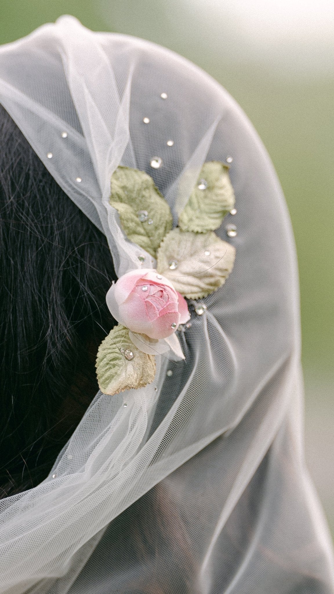 A close up of the Briar Juliet Cap Wedding Veil with sheer tulle, dotted crystals, and a rose with leaves sits atop a bride's head. 