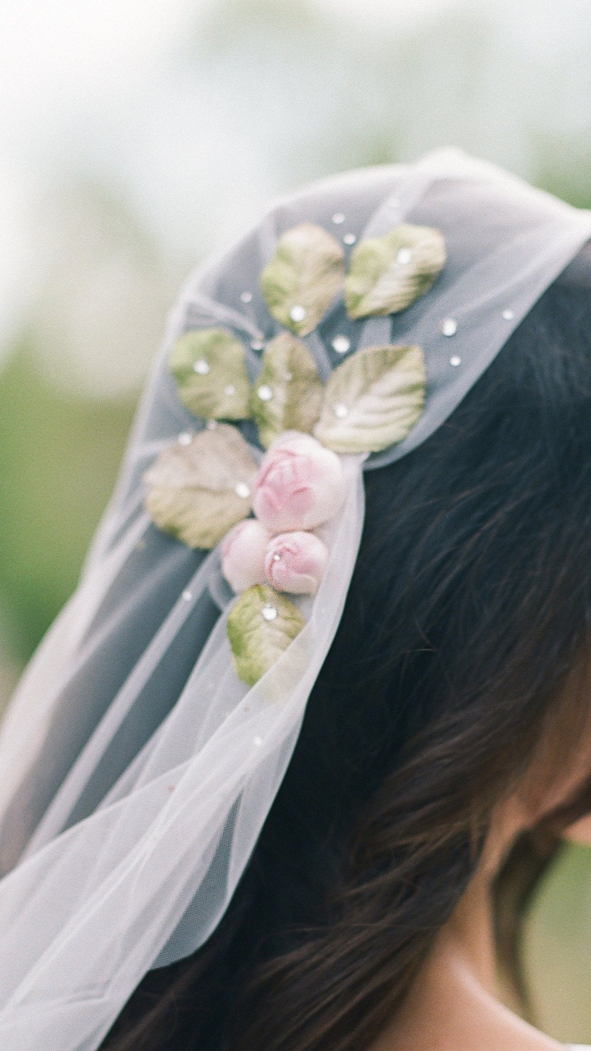 A close up of the Briar Juliet Cap Wedding Veil with sheer tulle, dotted crystals, and a rose with leaves sits atop a bride's head. 