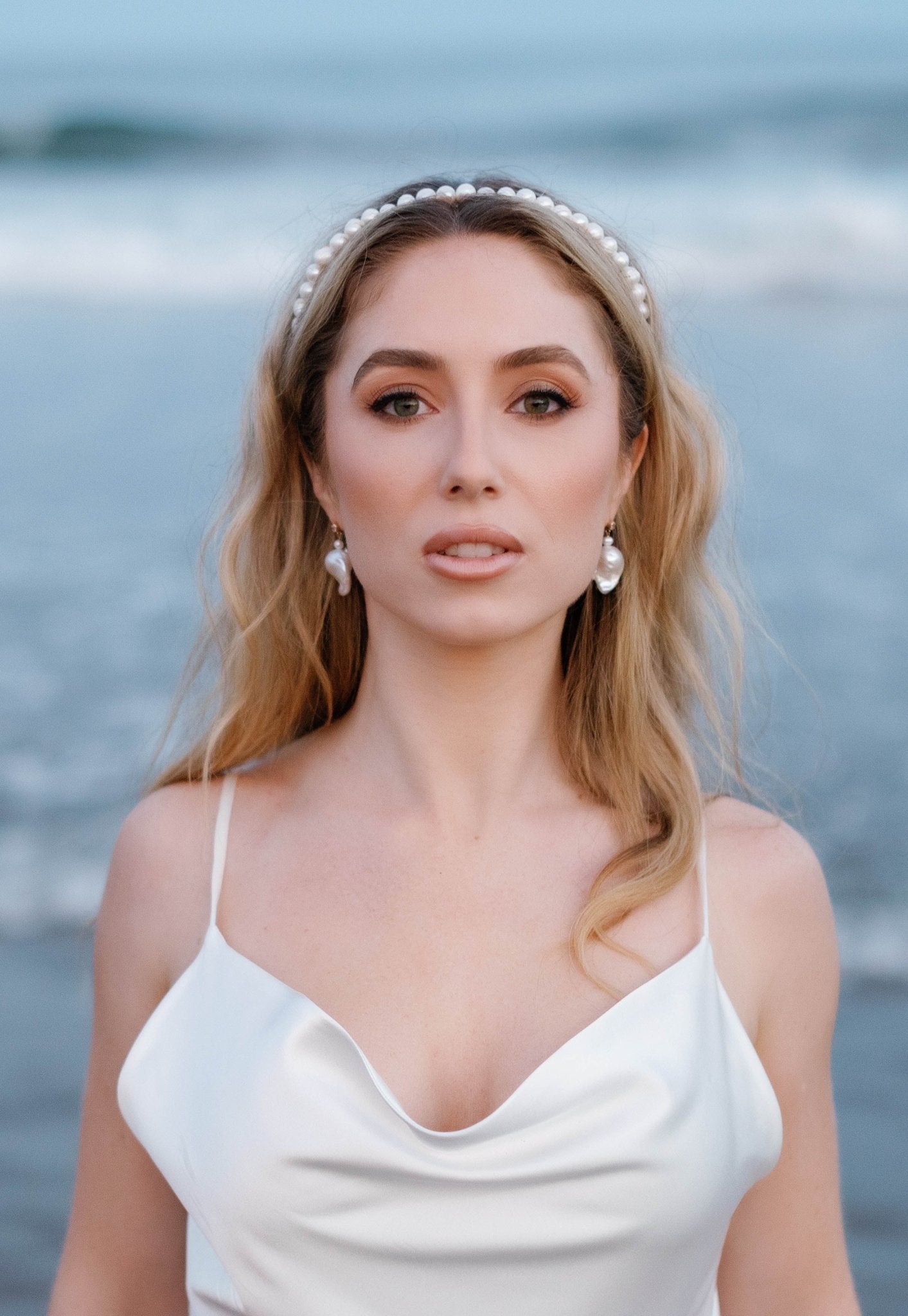 An beach-going bride is backed by the ocean as she wears a silky bridal gown, baroque pearl earrings, and her bubbled freshwater pearl headband. 