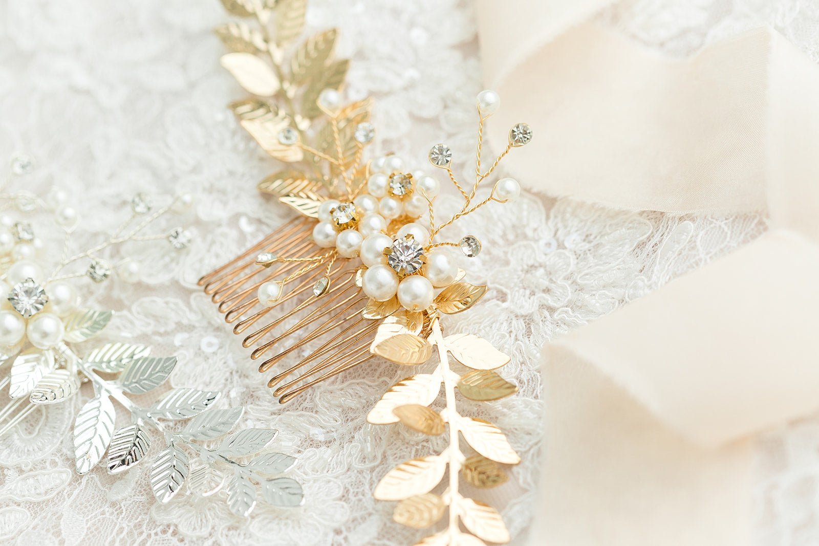 A bridal hair comb with freshwater pearls, crystals, and golden leaves lays on a length of lace above a silver one.  