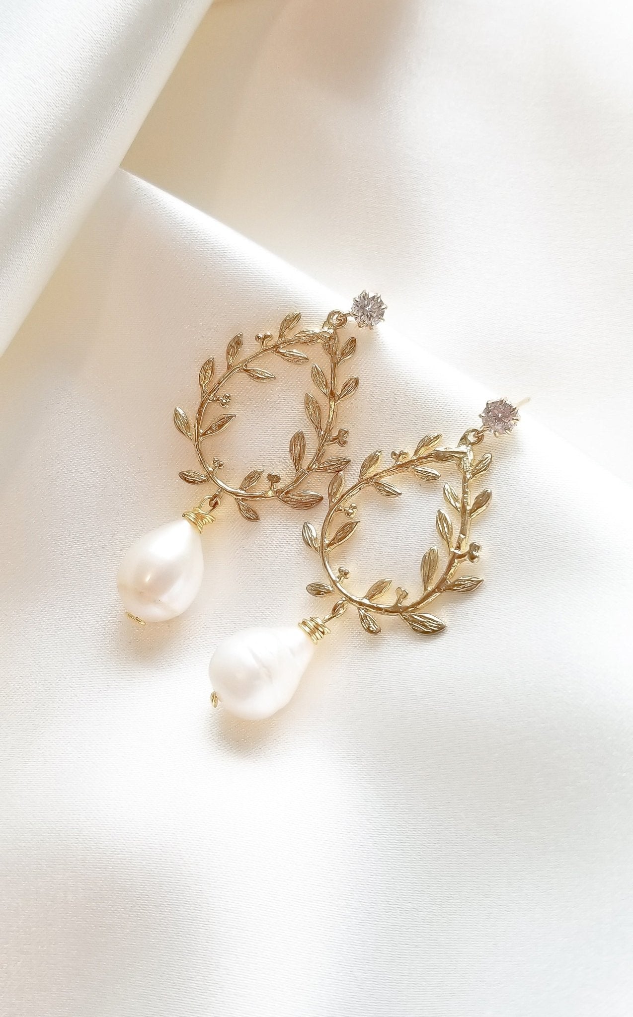 A pair of gold-filled, laurel leaf bridal earrings with freshwater pearl drops and crystal studs on white fabric background. Pearl Bridal jewelry from Canada, organic pearl jewelry, & statement bridal earrings, handmade by Canadian jewelry brand 