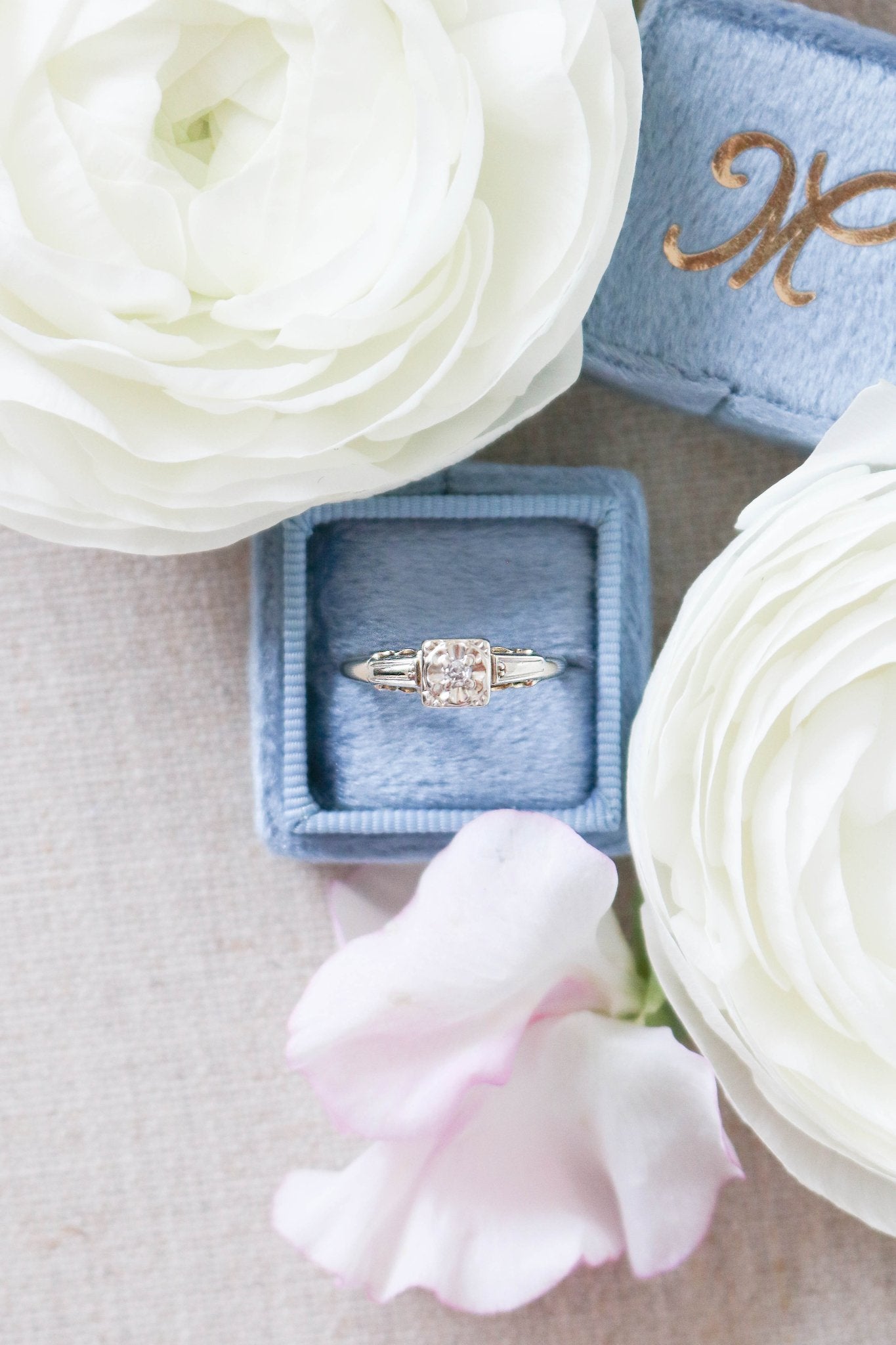 A gorgeous Gold Vintage Diamond Engagement Ring displayed in a powder blue ring box surrounded by white roses. 