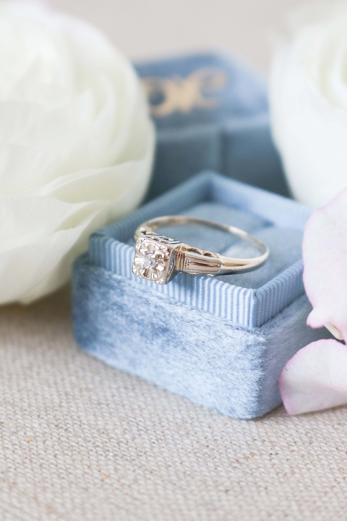 A Cayley Diamond Solitaire Vintage Engagement Ring in 14ct Gold, Art Deco Style from the 1930s sits atop a powder blue ring box. 