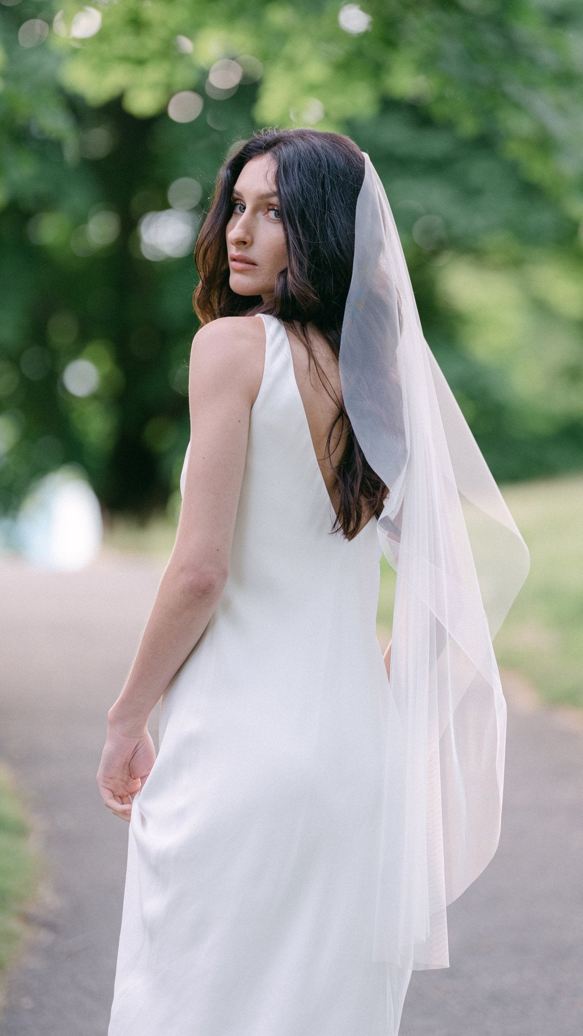 A bride all in white looks back at the camera while wearing a simple sheer and light wedding veil. 