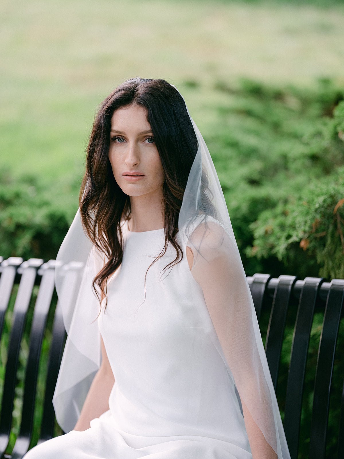 Sitting on a bench, a bride looks on as her sheer light bridal veil drapes around her. 