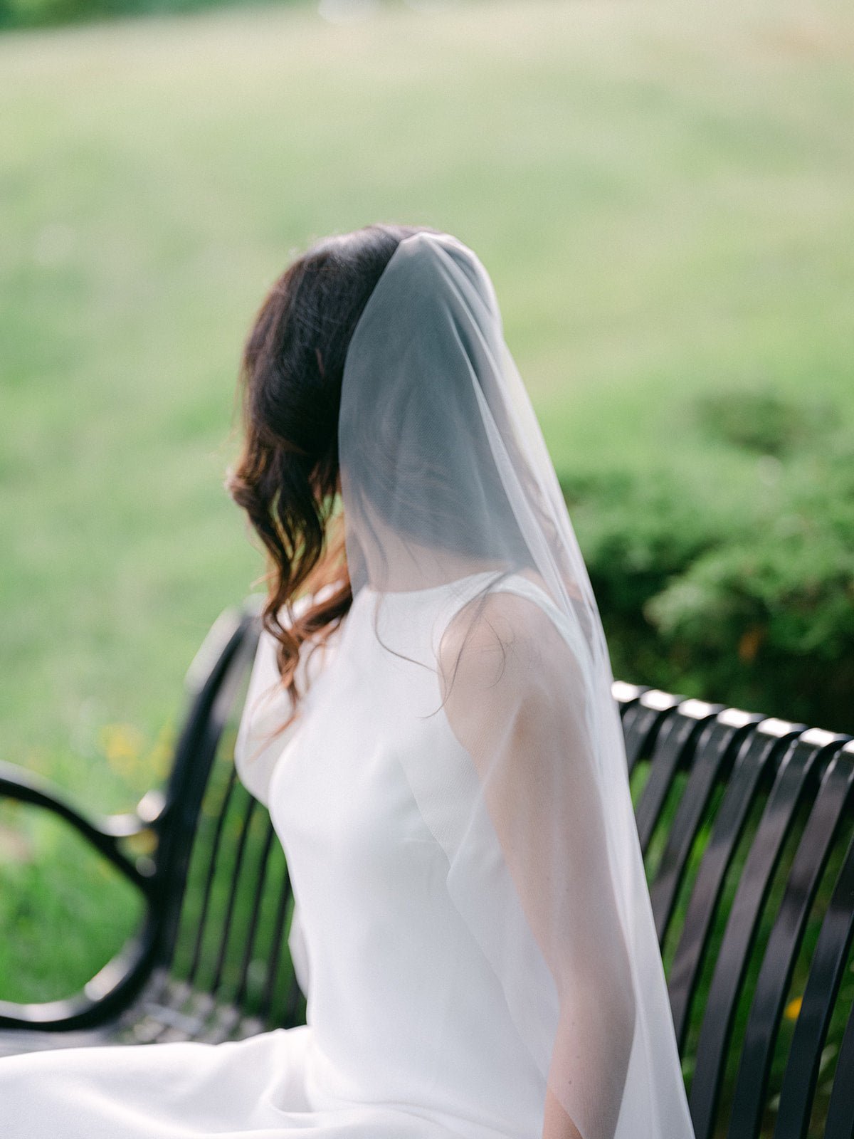 Sitting on a bench, a bride looks away as her sheer light bridal veil drapes around her. 