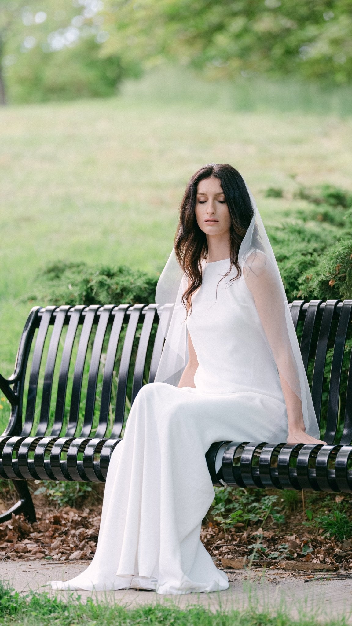 With eyes closed, a bride sits on a bench in a white wedding dress and a sheer light bridal veil. 