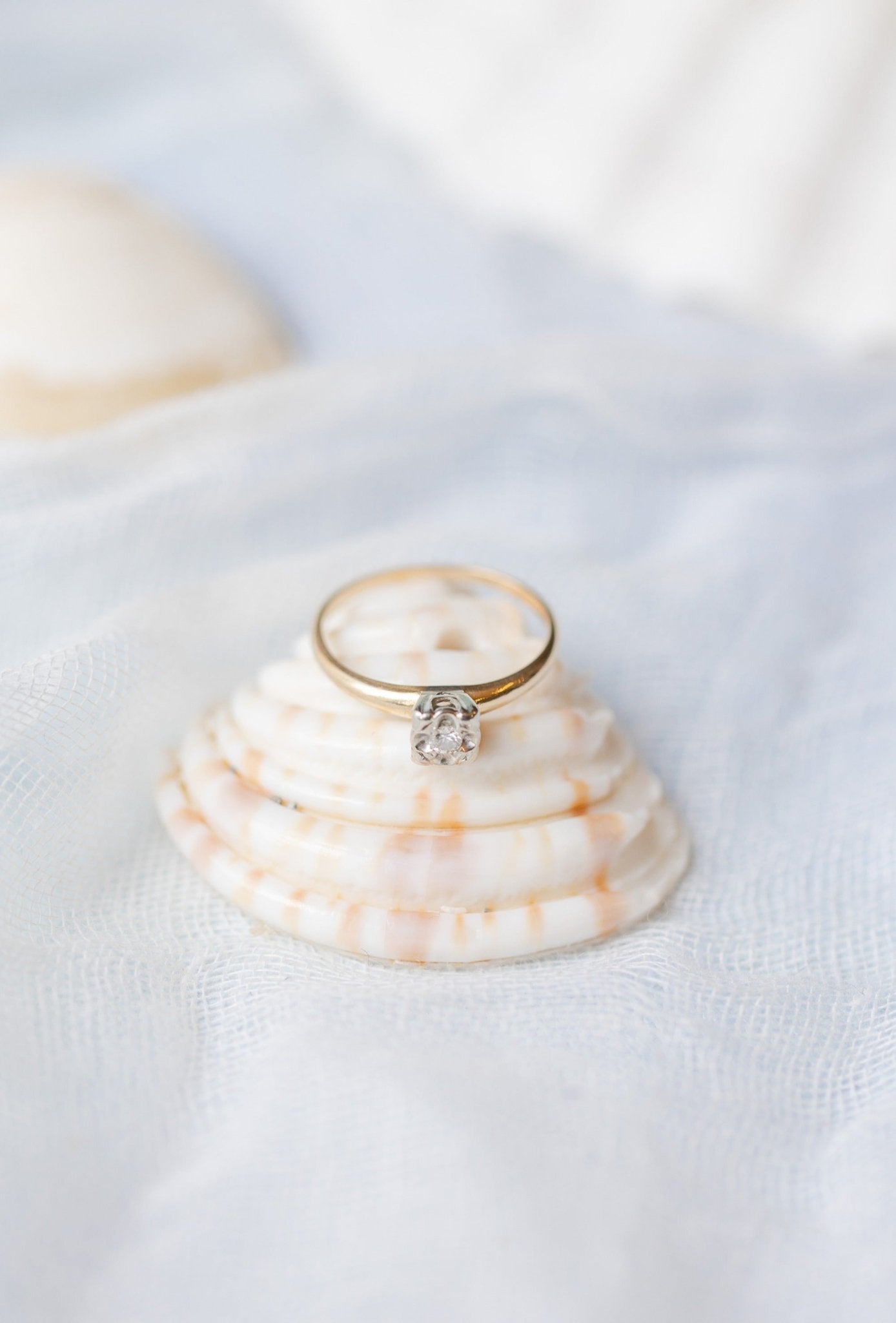 The Claire Vintage Diamond Solitaire Engagement Ring in 14k Yellow and White Gold sits on top of a shell against a white background. 