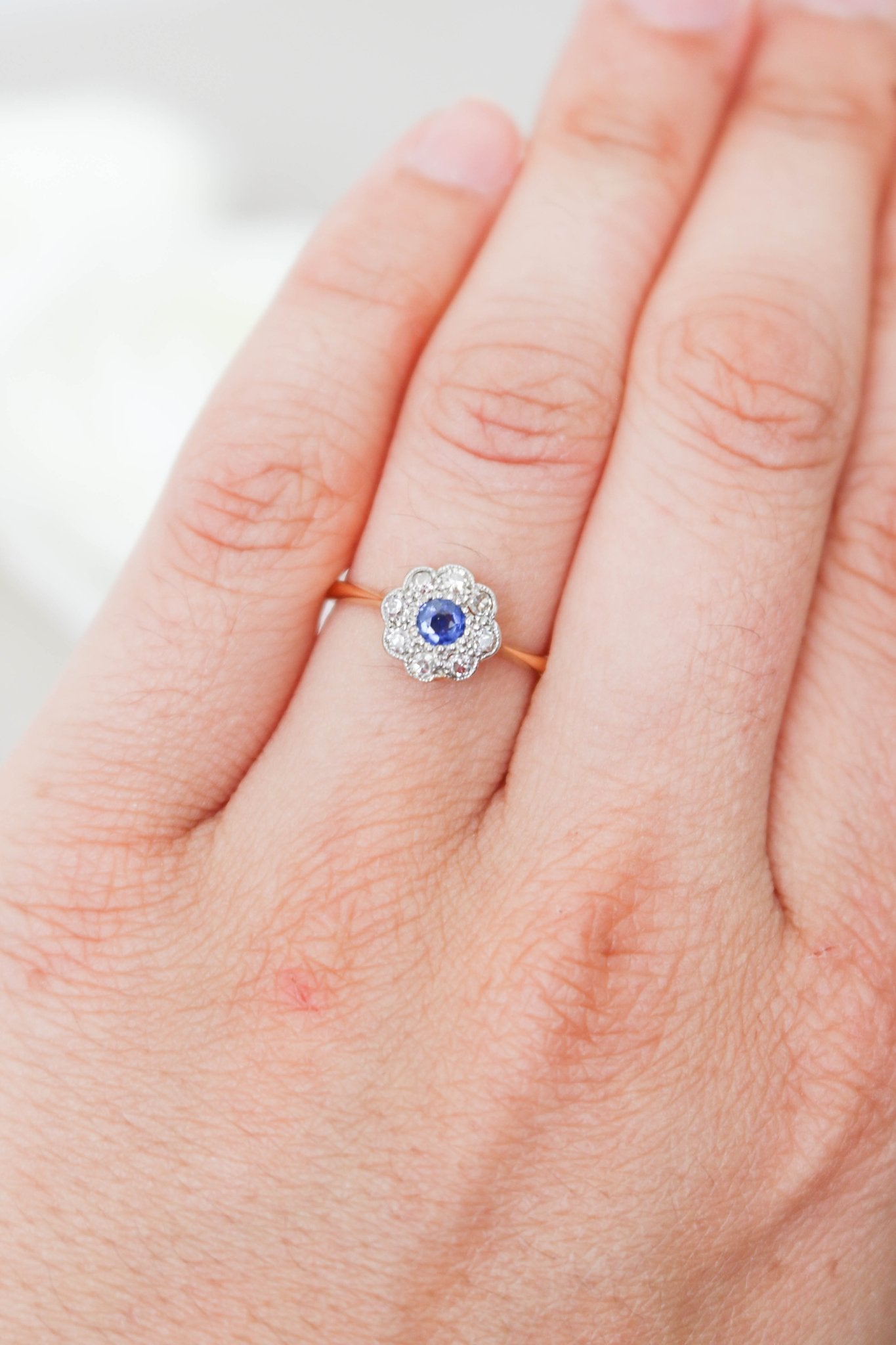 An 18k Yellow and White Gold flower design Vintage Engagement Ring with Sapphire and Diamond displayed on a ring finger. 