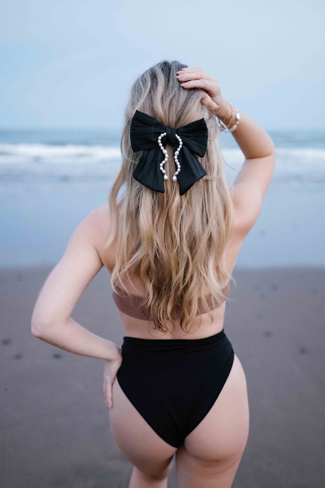 Beach babe in bathing suit from behind showcasing her satin black Delphine Bow with freshwater pearls.