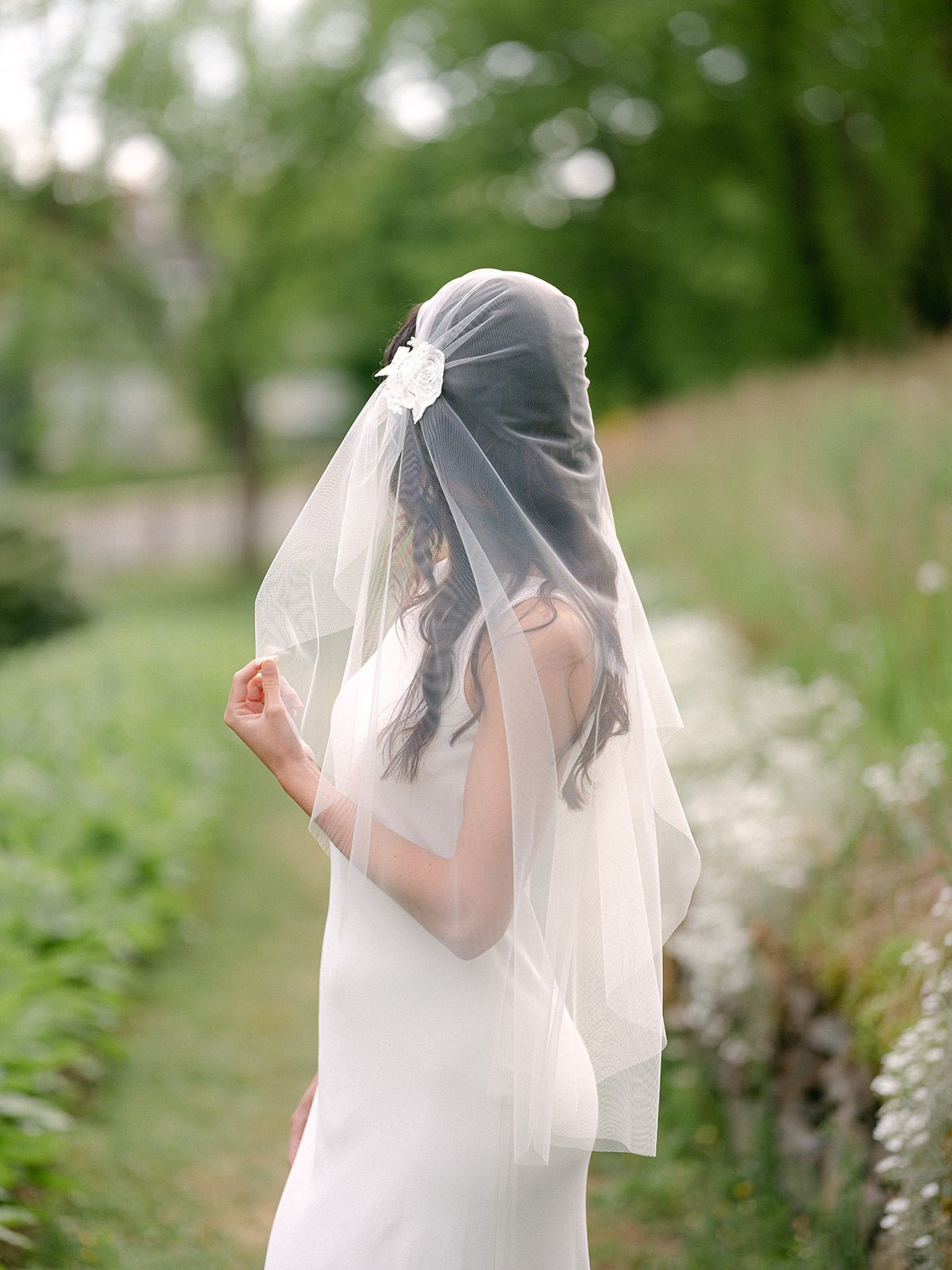 A bride looking away while holding up part of her sheer tulle wedding veil amongst greenery. 