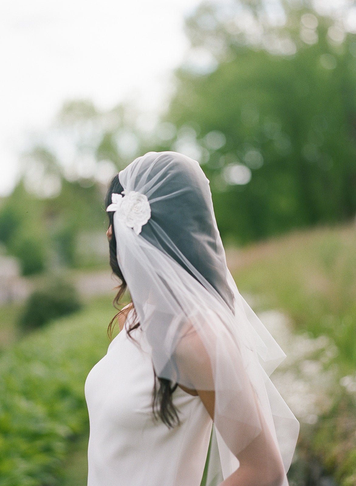 A bride looking away while wearing a sheer tulle wedding veil amongst greenery. 