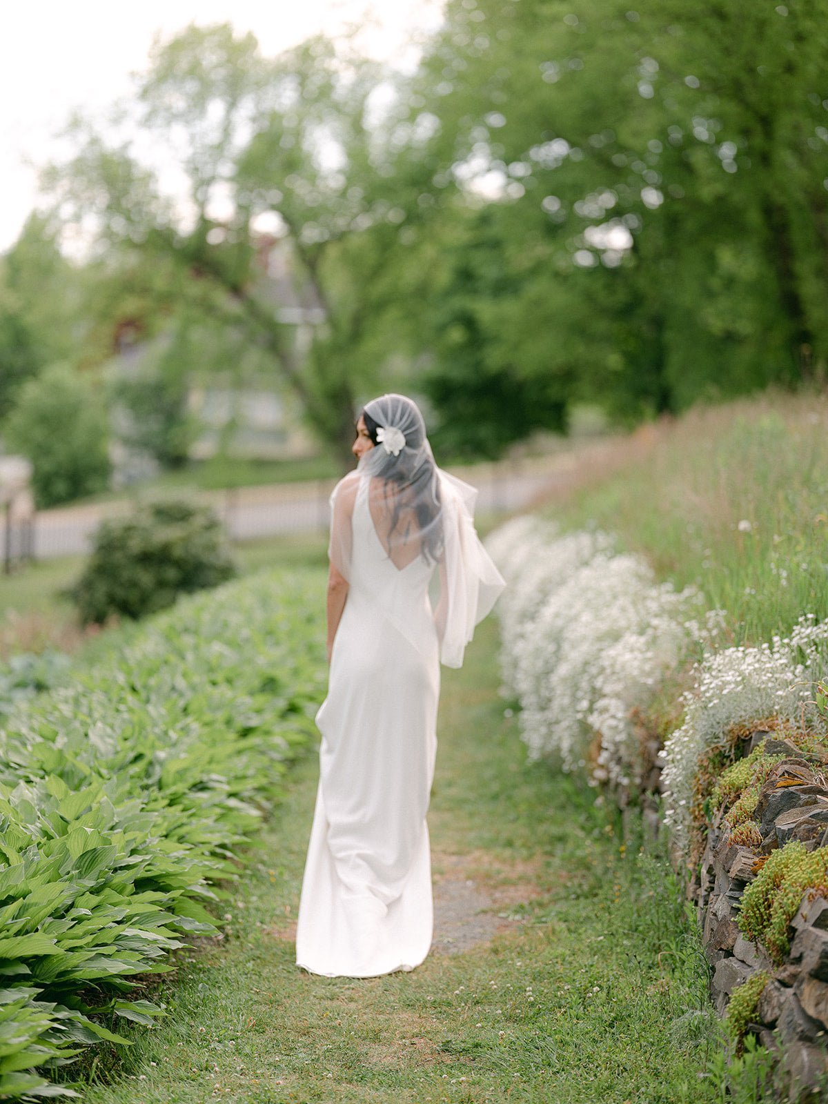 A bride surrounded by foliage glances to the side in her bunched wedding veil with embroidered flowers on the sides. 