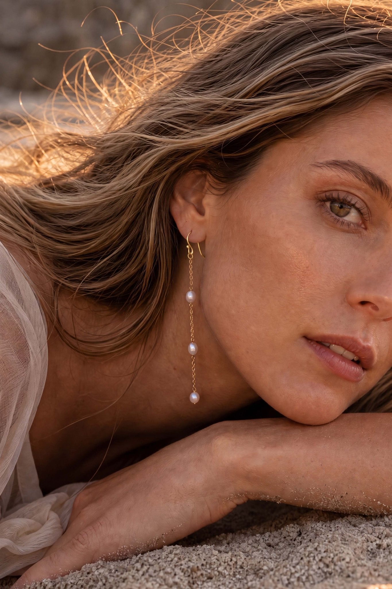 A close-up lifestyle shot of a woman lying on sand, wearing a delicate gold dangle freshwater pearl earrings in 14k gold filled.
