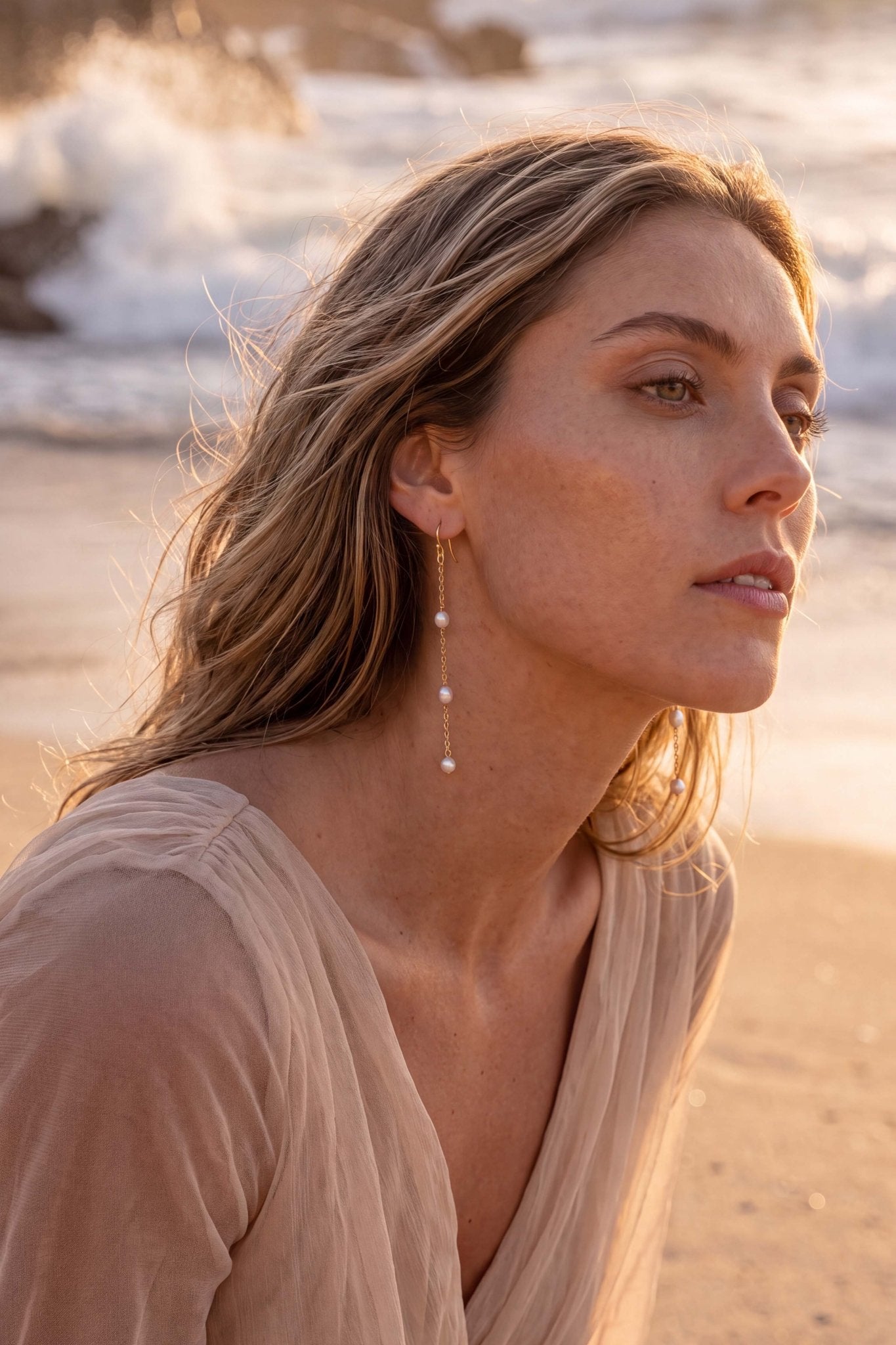 A woman with blonde hair wearing a chiffon dress and long, dangling 14k gold filled pearl-earrings, photographed outdoors on a sandy beach with ocean waves in the background.