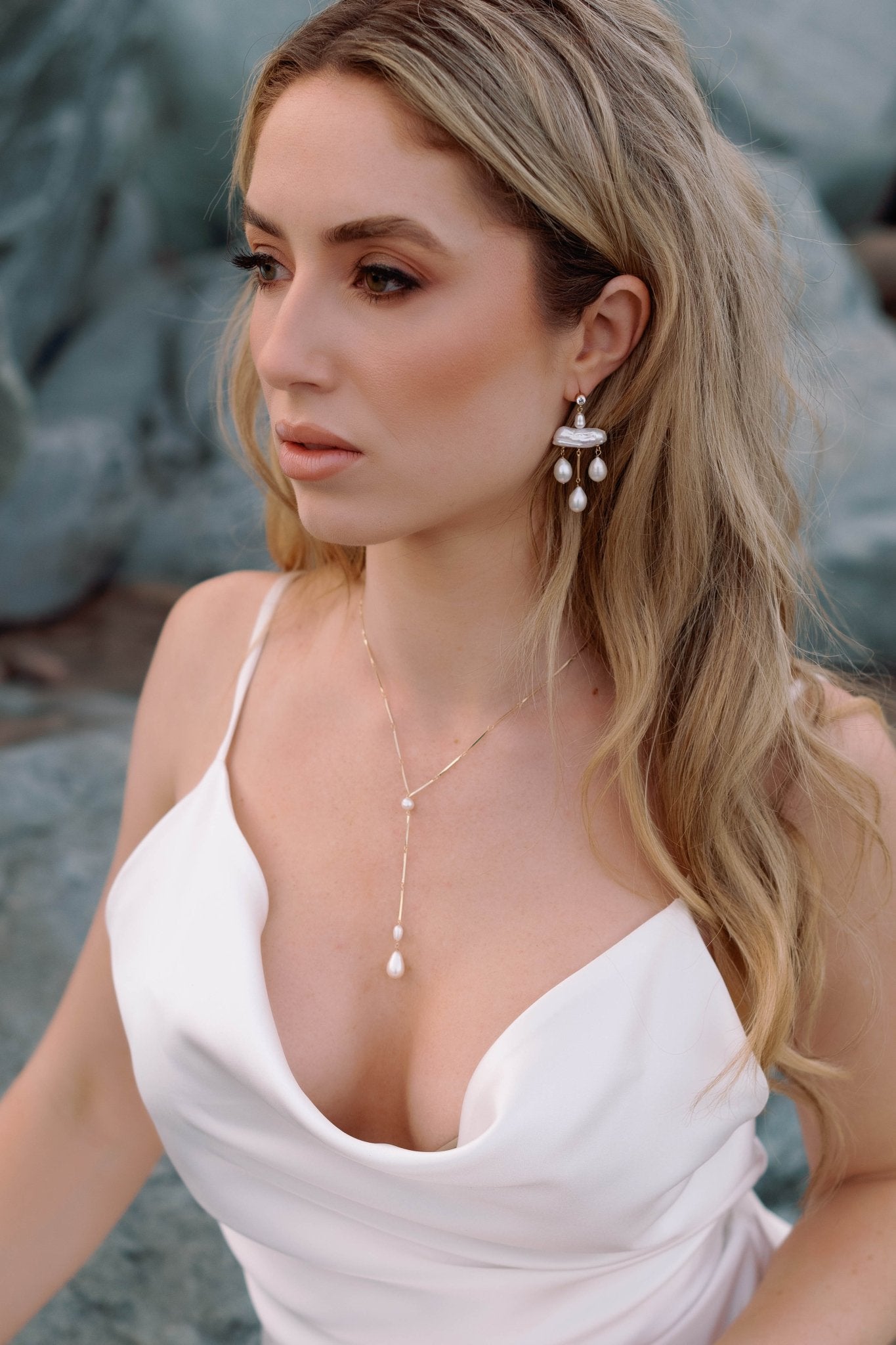 Inquisitive beach bride gazes ahead dressed in a satin custom wedding dress. She is draped in subtle luxury with a gold-filled pearl drop necklace and pearl chandelier earrings.