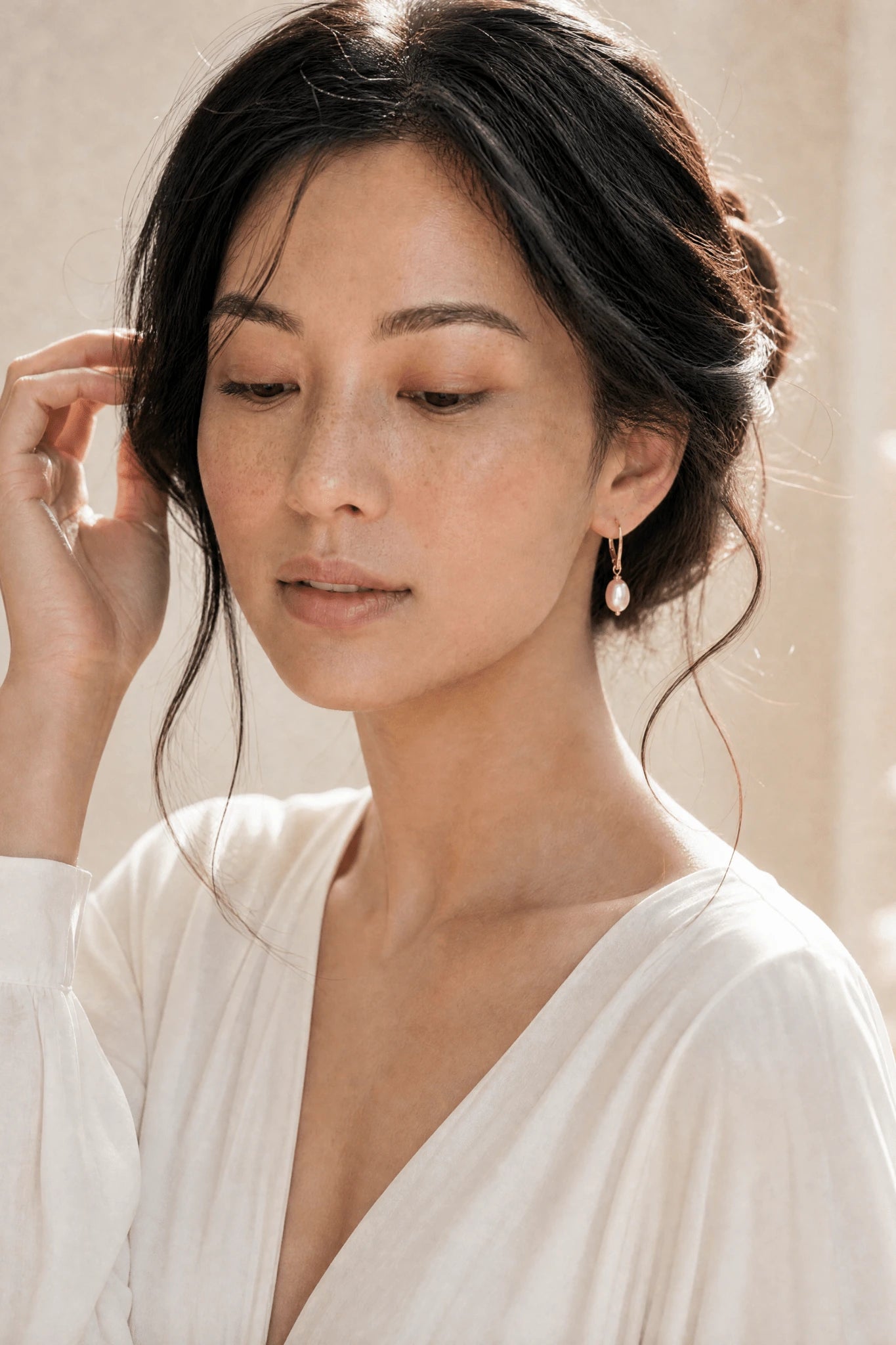Filipino woman in white dress looking down while wearing gold blush pearl earrings against neutral background. Handmade pearl jewelry by Canadian Jewelry artist
