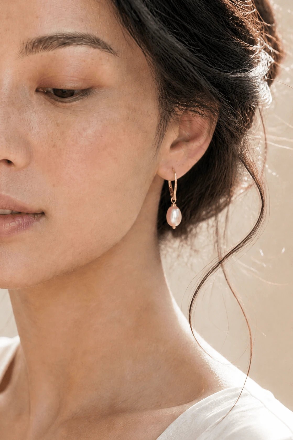 Close up of BIPOC model with gold filled leverback earring with fresh water blush pearl drop against neutral background. 