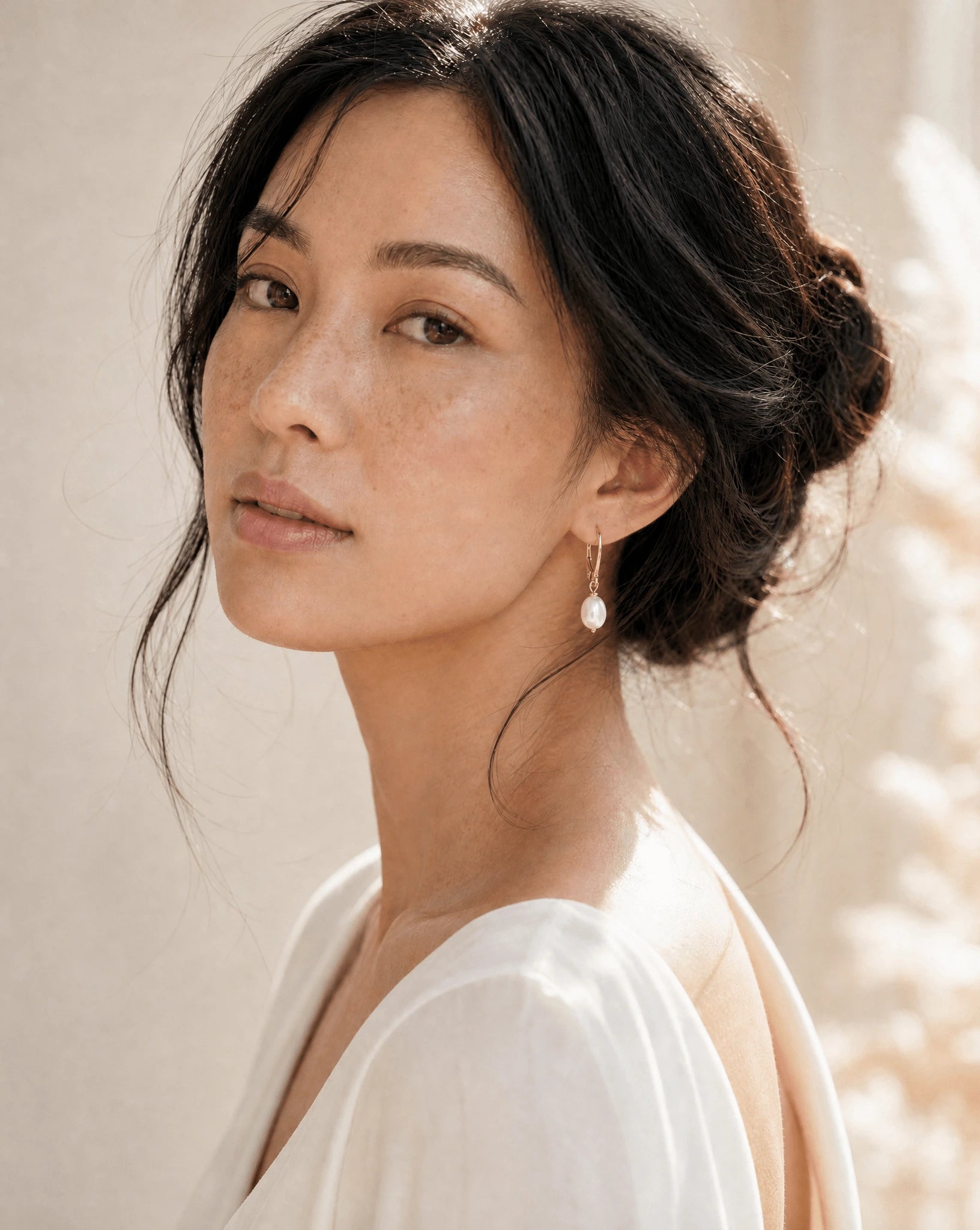 Filipino woman with messy bun in white wearing gold fresh water pearl earrings with leverback hooks against neutral background