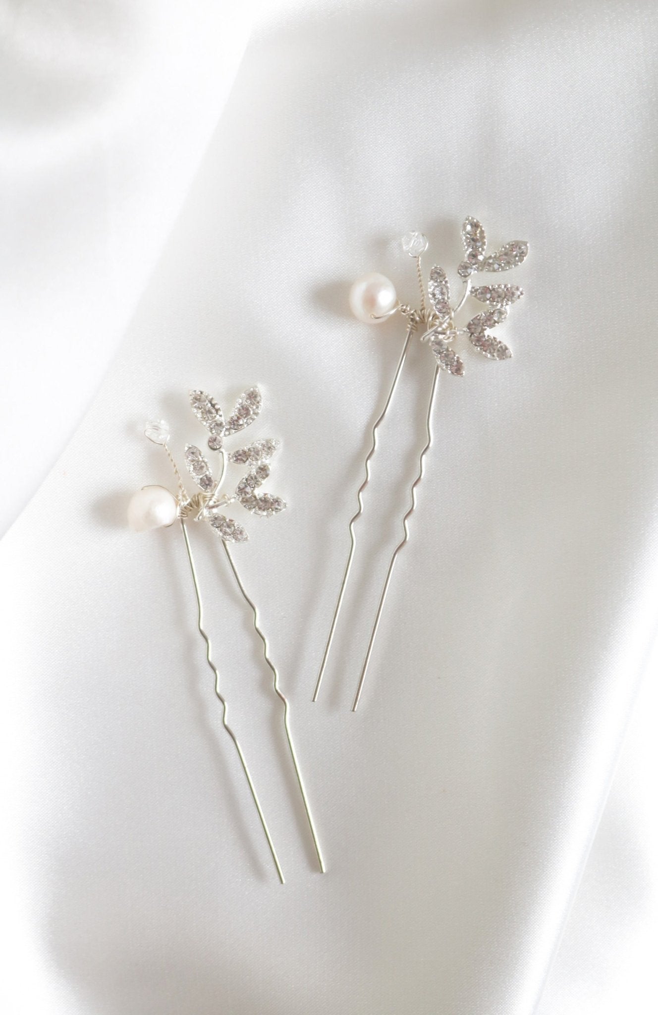 Two silver bridal hairpins with leaf-shaped settings adorned with sparkling crystals and fresh water pearls placed on a white fabric surface. Bridal hair accessories & pearl bridal accessories handmade in Canada by Canadian jewelry designer
