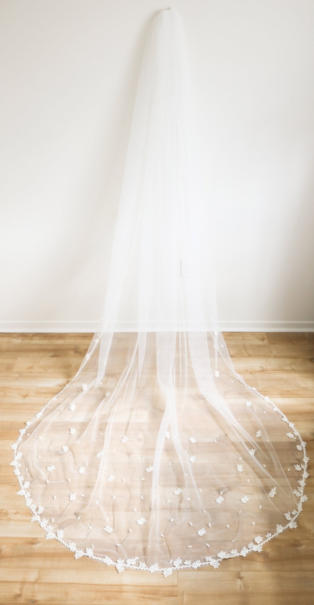 A Sheer bridal veil hangs on a wall; its embroidered flowers grace the edges of the tulle that flows along the wooden floor. 