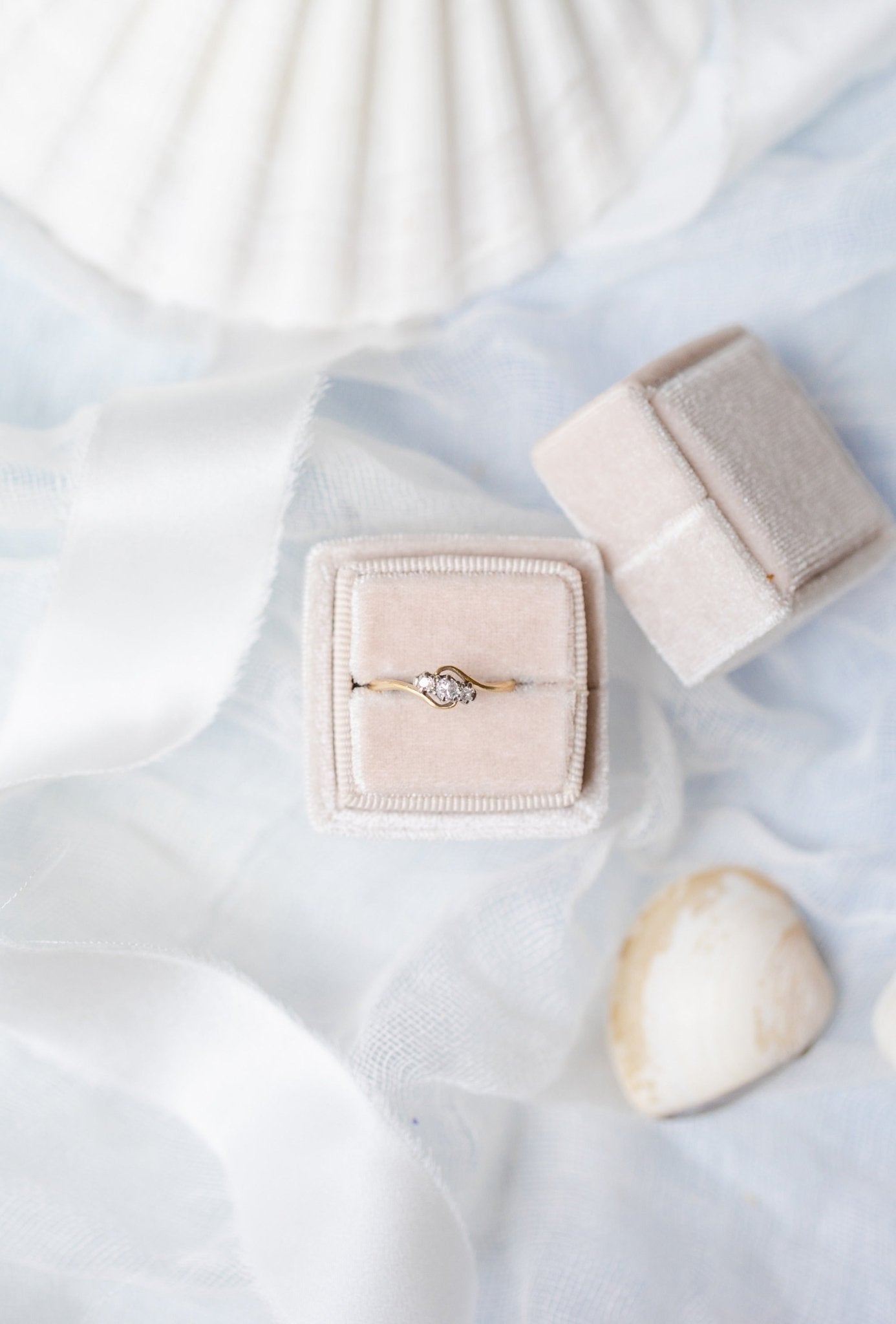 An Emilia Trilogy Diamond Engagement Ring in 18ct Gold and Platinum, c.1900 England sitting in a ring box against a white backdrop with seashells. 