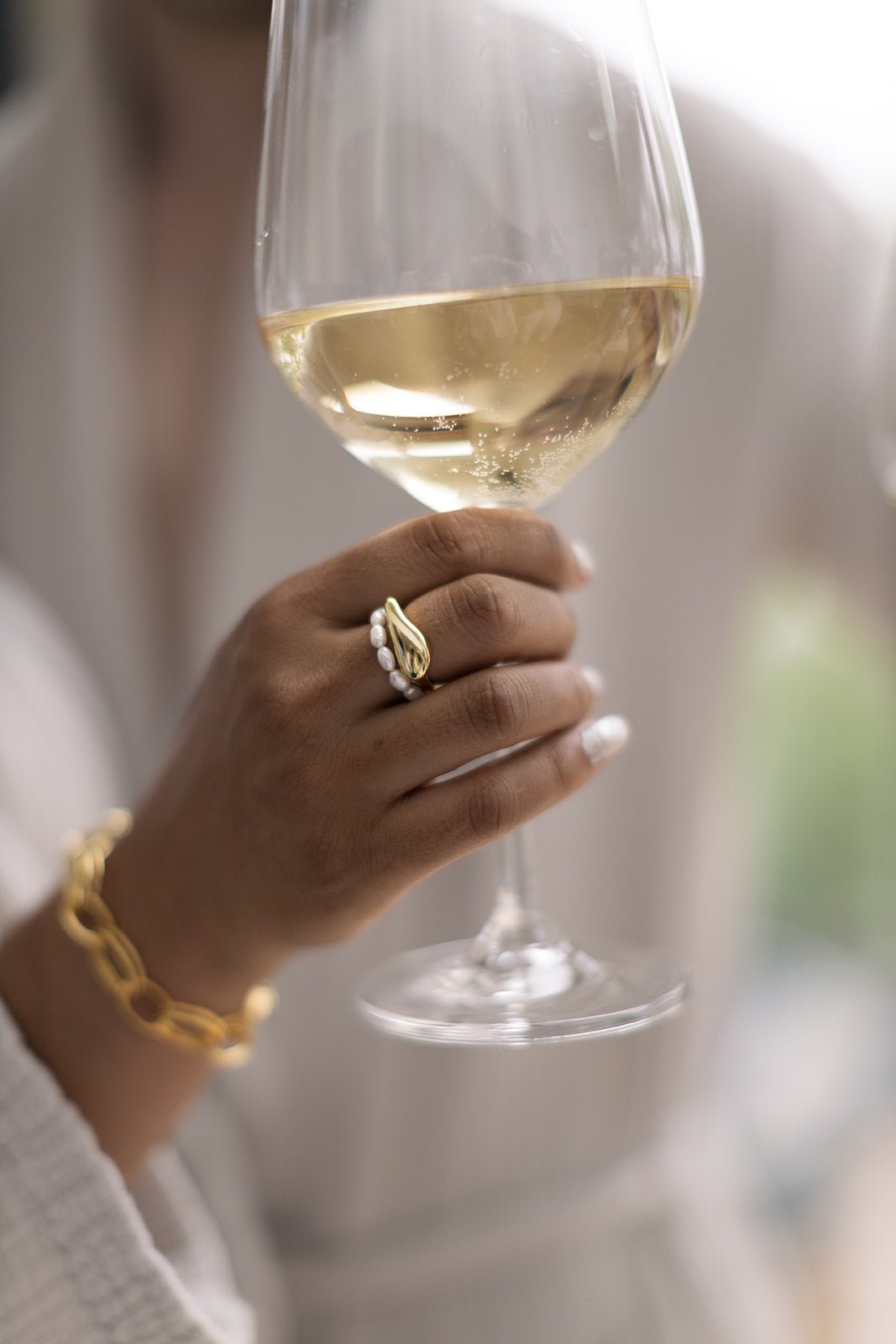 Hand holding a glass of white wine and showcasing stacked rings with the Everyday Freshwater Pearl Ring and the Essence Figure Gold-Filled Ring. Gold-filled jewelry Canada. Canadian jewelry designs.