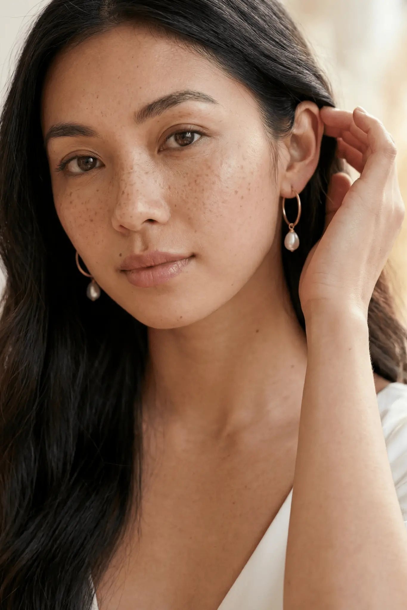Filipina woman tucking hair behind her ear wearing gold filled earrings with freshwater pearls on a neutral background. Canadian Pearl jewelry & gold filled jewelry handmade by BIPOC jewelry designer