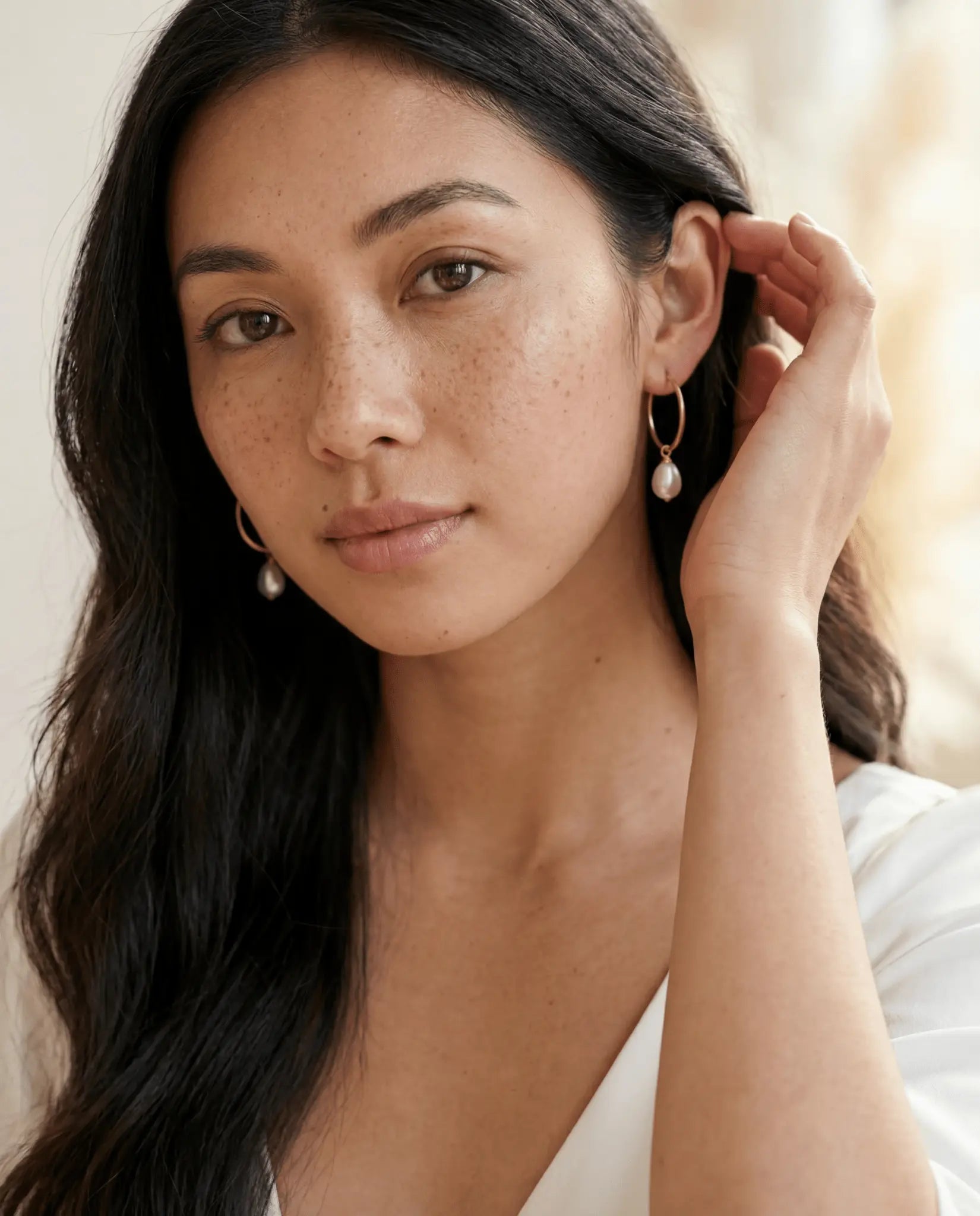Filipina woman tucking hair behind her ear wearing gold filled earrings with freshwater pearls on a neutral background. Canadian Pearl jewelry & gold filled jewelry handmade by BIPOC jewelry designer