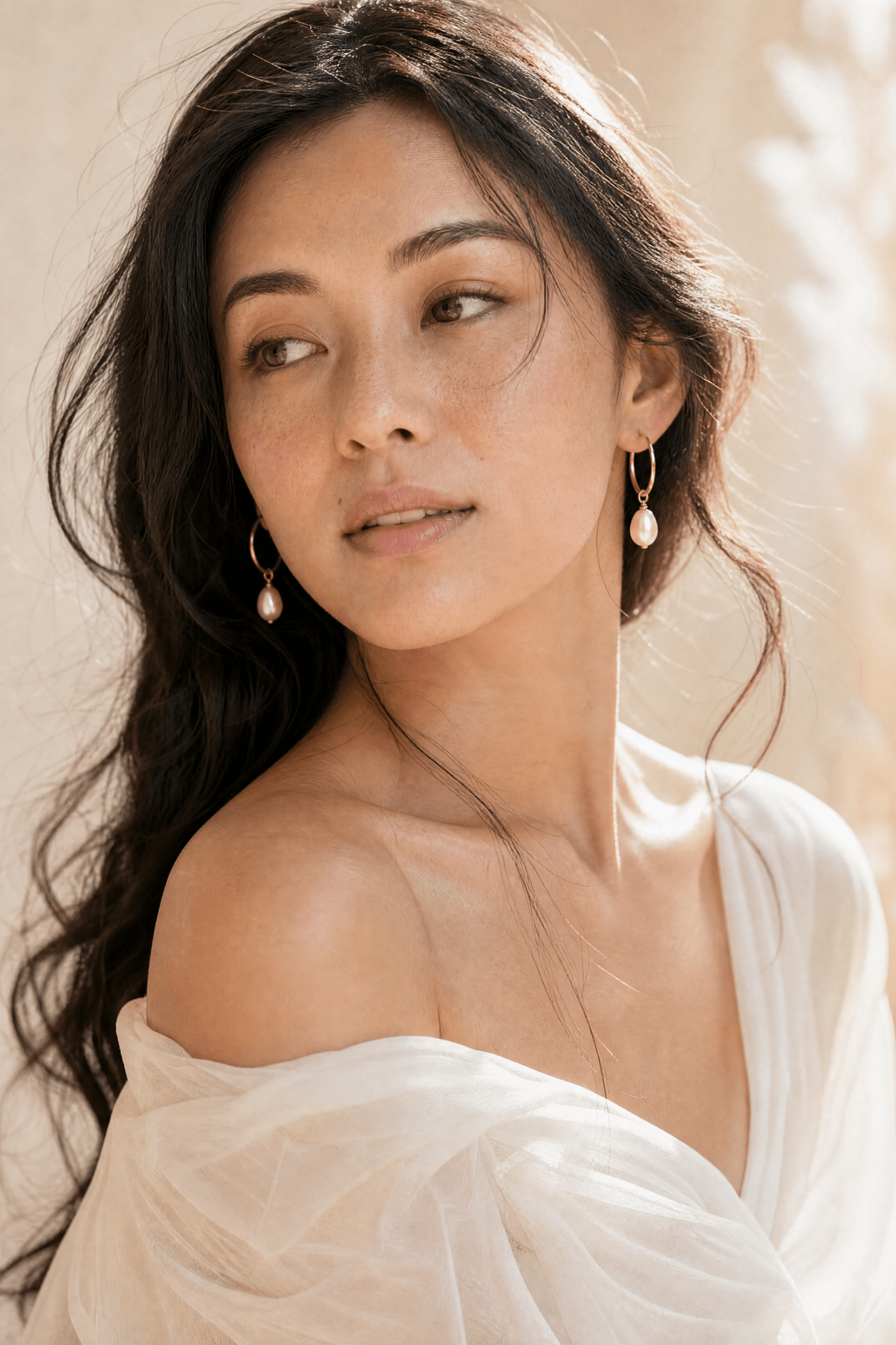 Filipino woman with long dark hair wearing a white off-shoulder top and gold blush freshwater pearl hoop earrings against a neutral background