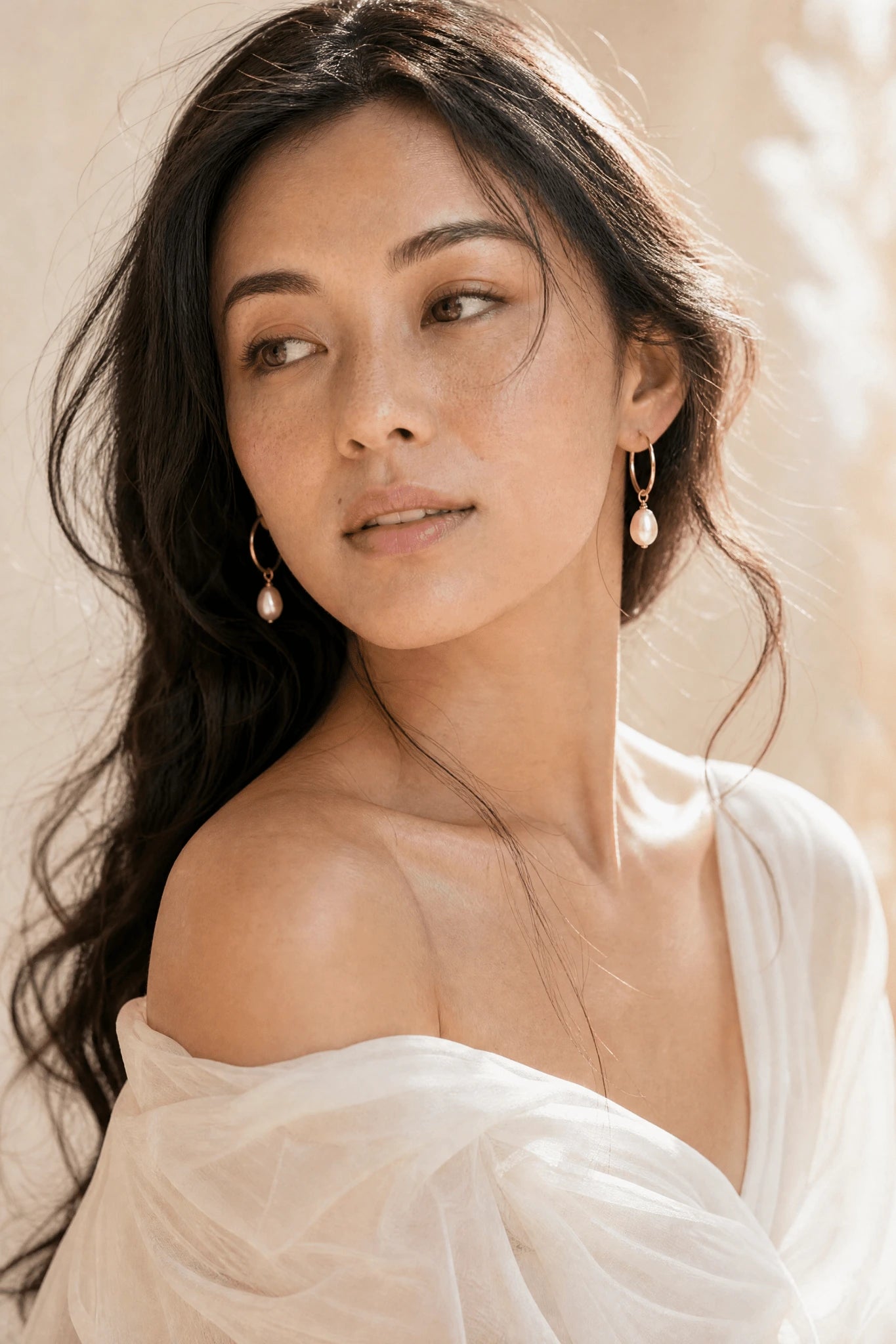 Filipino woman with long dark hair wearing a white off-shoulder top and gold blush freshwater pearl hoop earrings against a neutral background